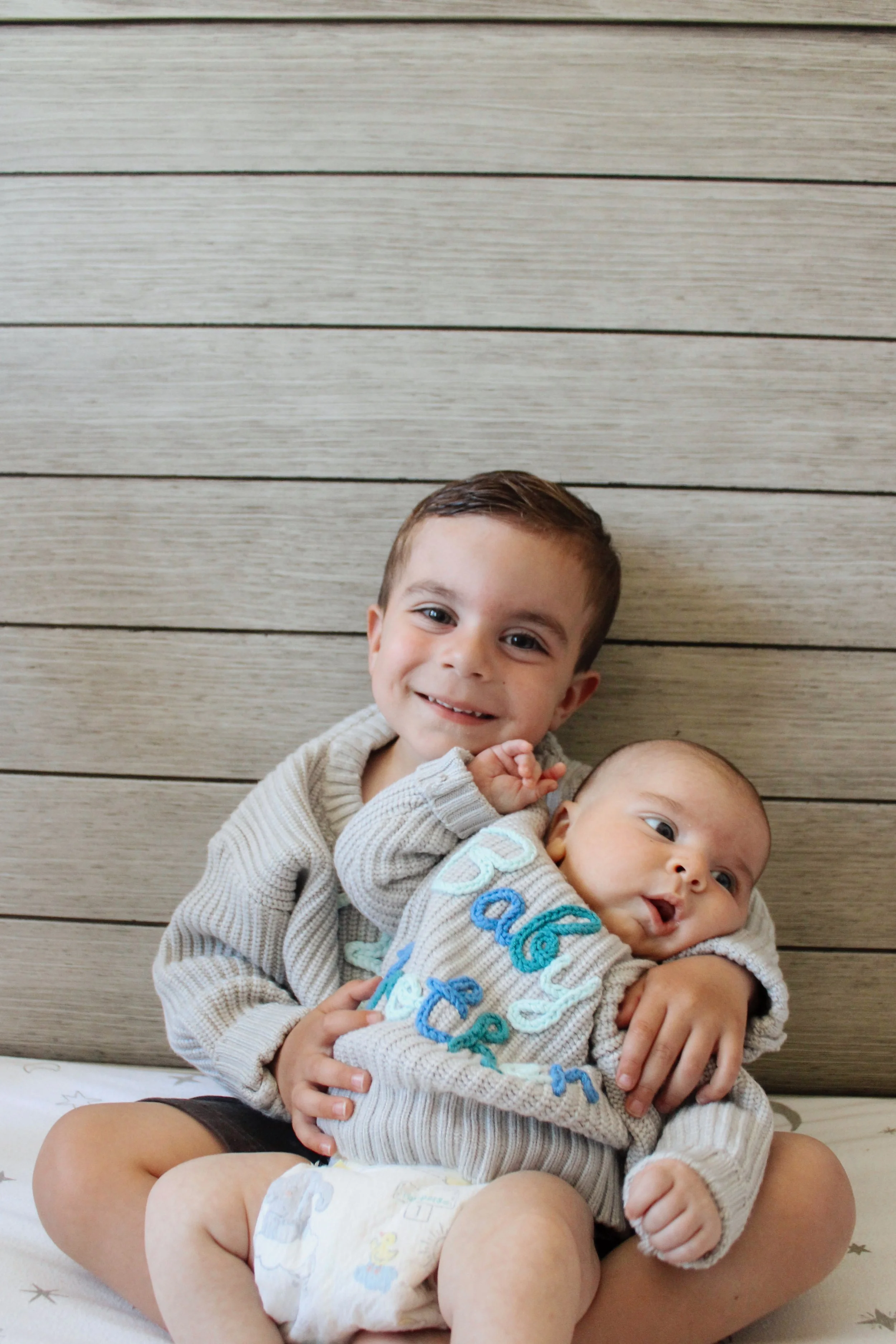 A young boy smiling while holding a baby who is looking off to the side.
