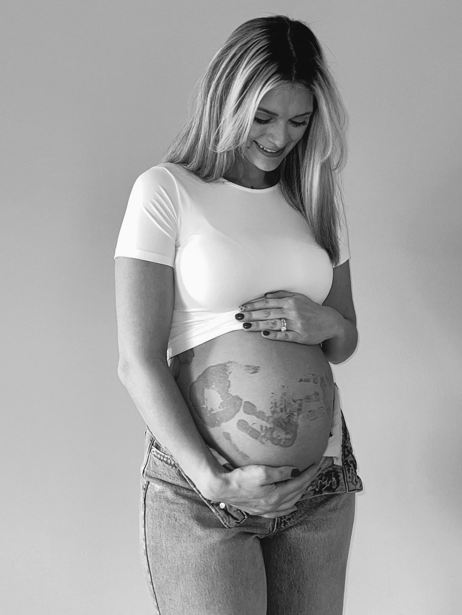 A pregnant woman with a tattoo on her belly, smiling and looking down, wearing a white t-shirt and jeans.