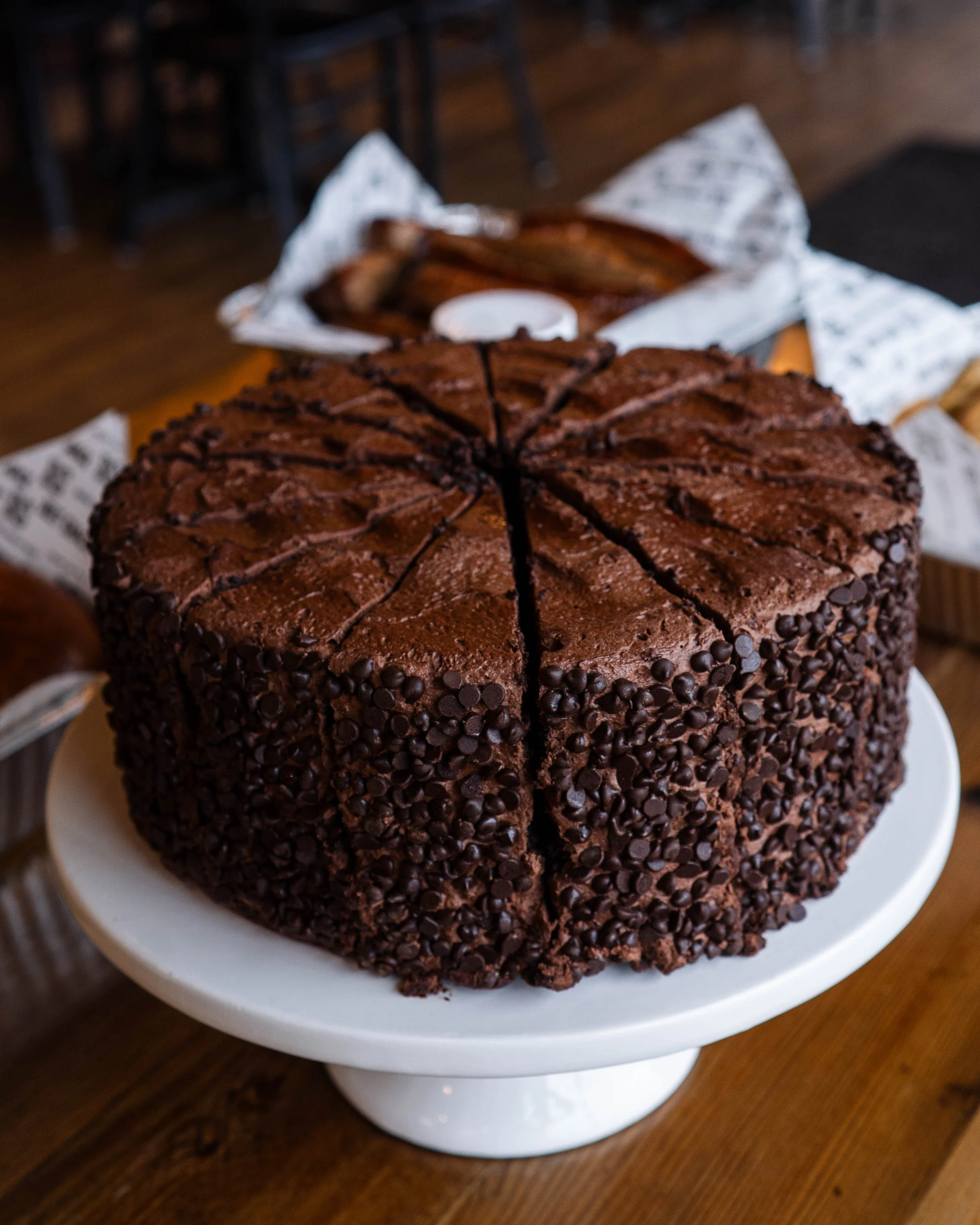 A large round chocolate cake covered in chocolate chips, sliced into pieces, on a white cake stand.