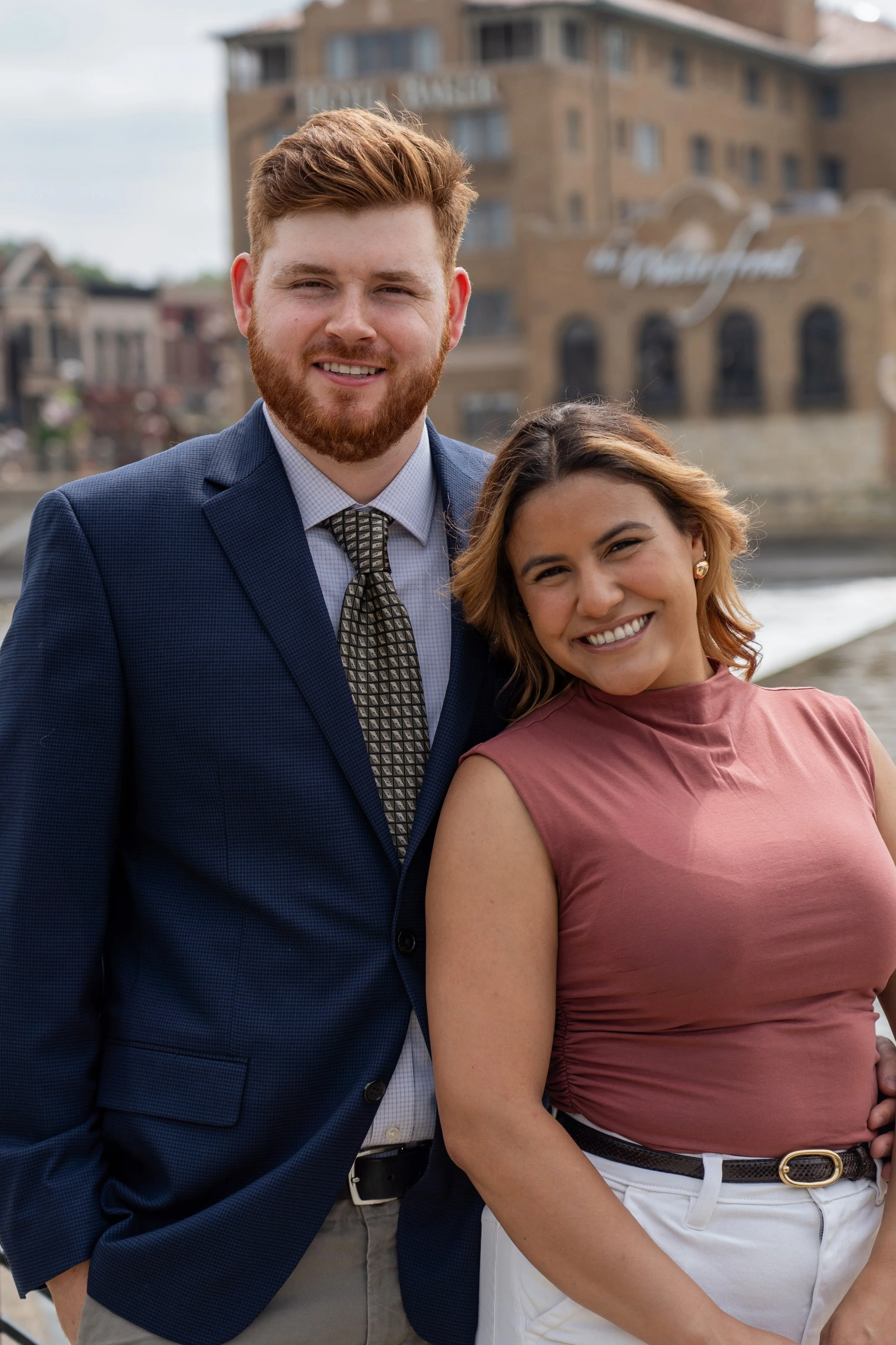 A man and woman smiling outdoors with city buildings in the background.