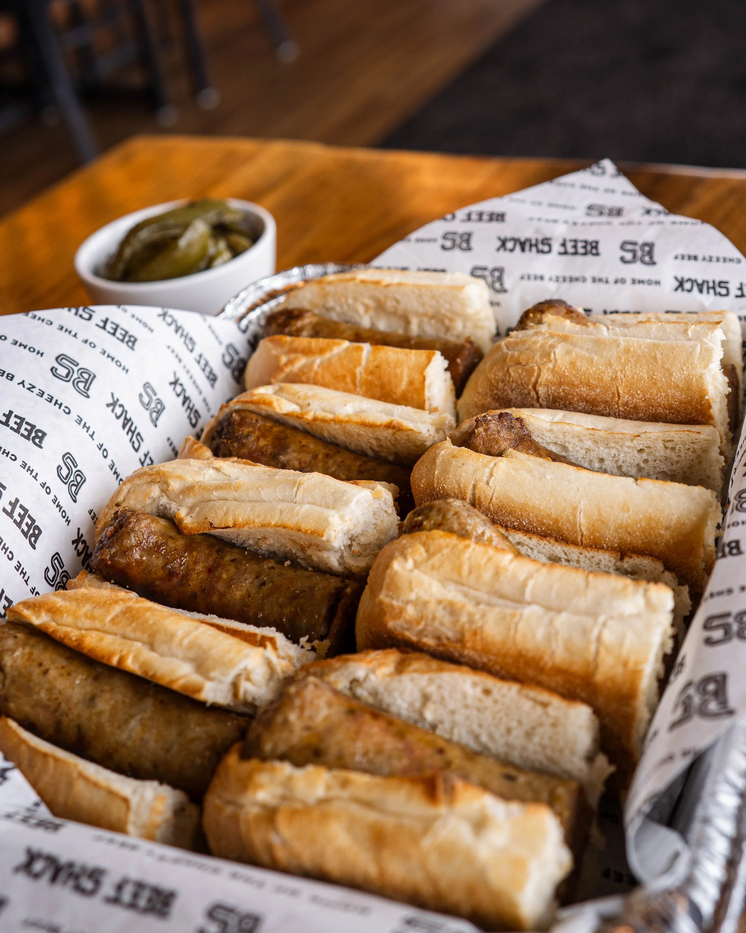 A basket filled with assorted hot dogs in buns, placed on parchment paper with 'BEE SHACK' printed on it. There is a small white bowl of pickles in the background.