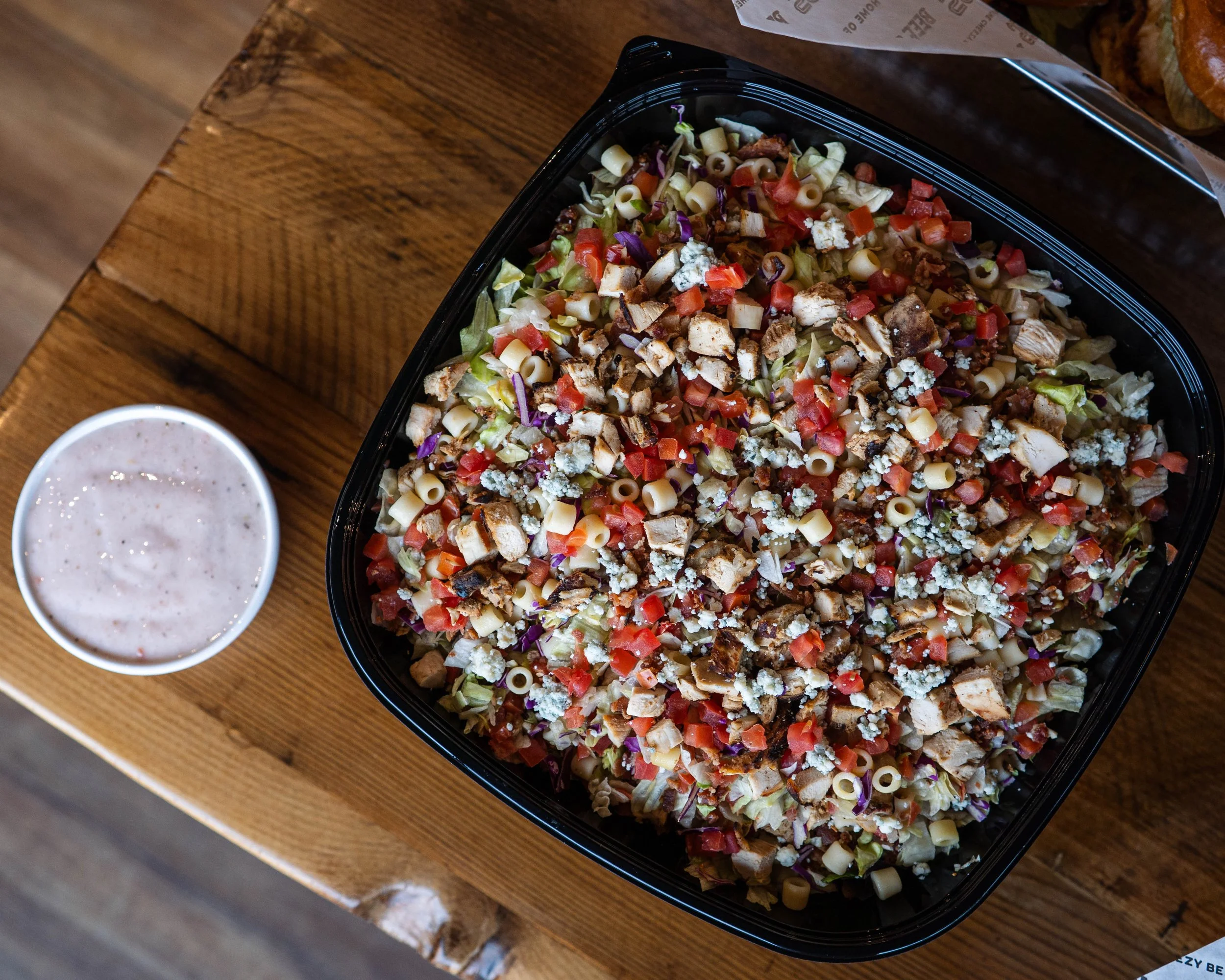 Large bowl of chopped salad with lettuce, tomatoes, pasta, grilled chicken, blue cheese crumbles, and chopped vegetables, with a side of creamy dressing on a wooden table.