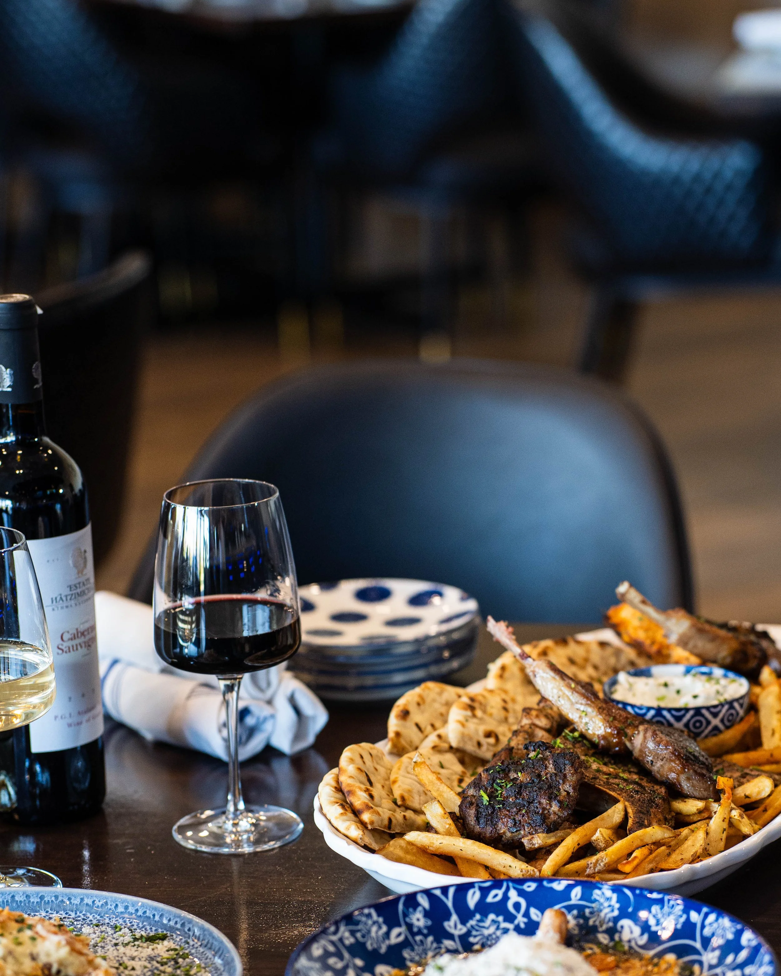 A dining table set with a glass of red wine, a bottle of wine, a plate of lamb chops with French fries, pita bread, and a bowl of dipping sauce, with a dark chair and blurred background.