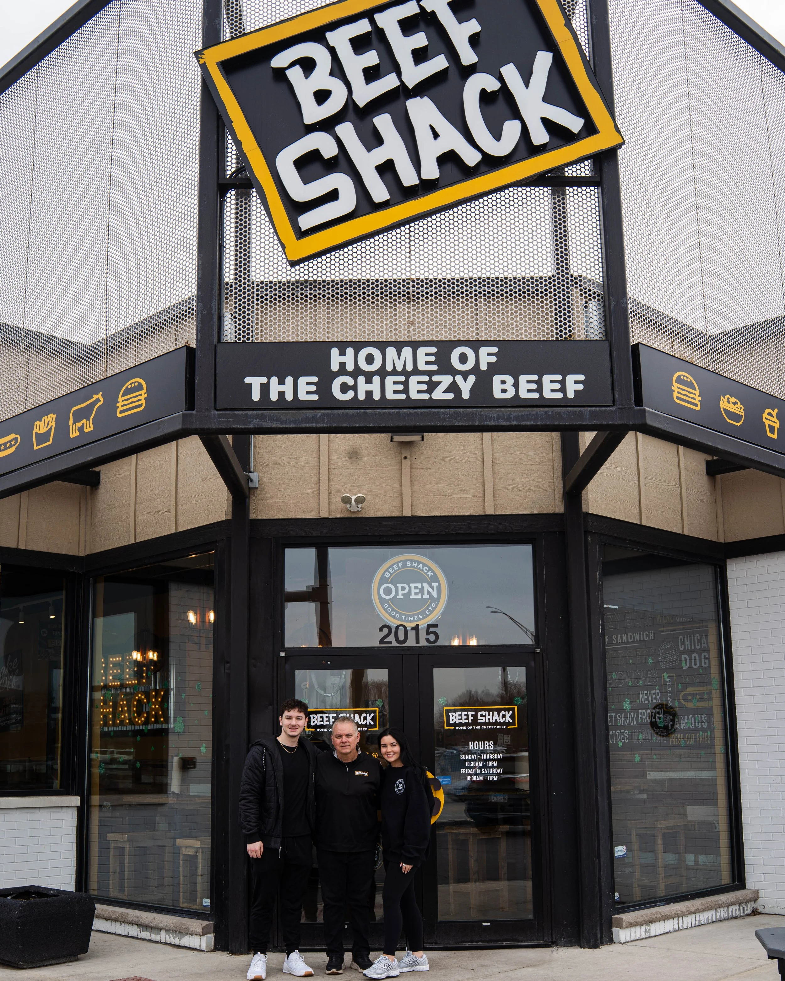 Exterior of a restaurant called Beef Shack, with a large sign reading 'Beef Shack' and a smaller sign below saying 'Home of the Cheezy Beef.' Three people stand in front of the door, smiling, with the restaurant's hours posted on the door, and it app