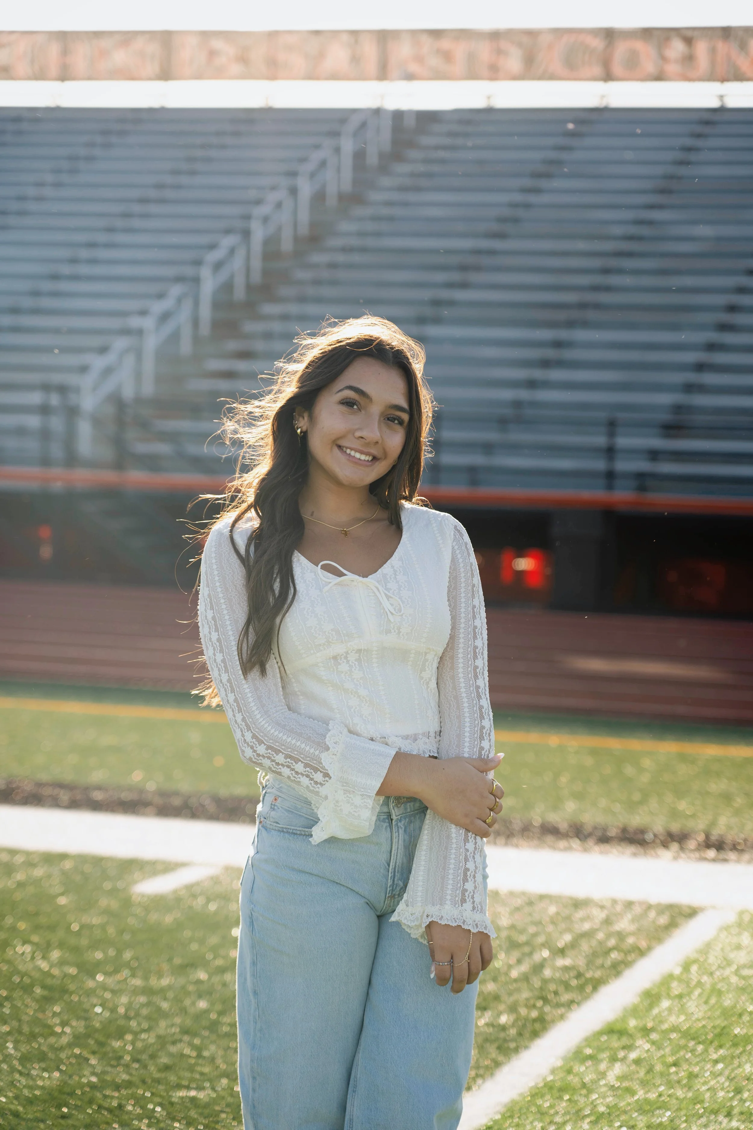 A young woman with long dark hair and a smile standing on a football field with bleachers in the background, wearing a white lace blouse and light blue jeans, illuminated by sunlight.