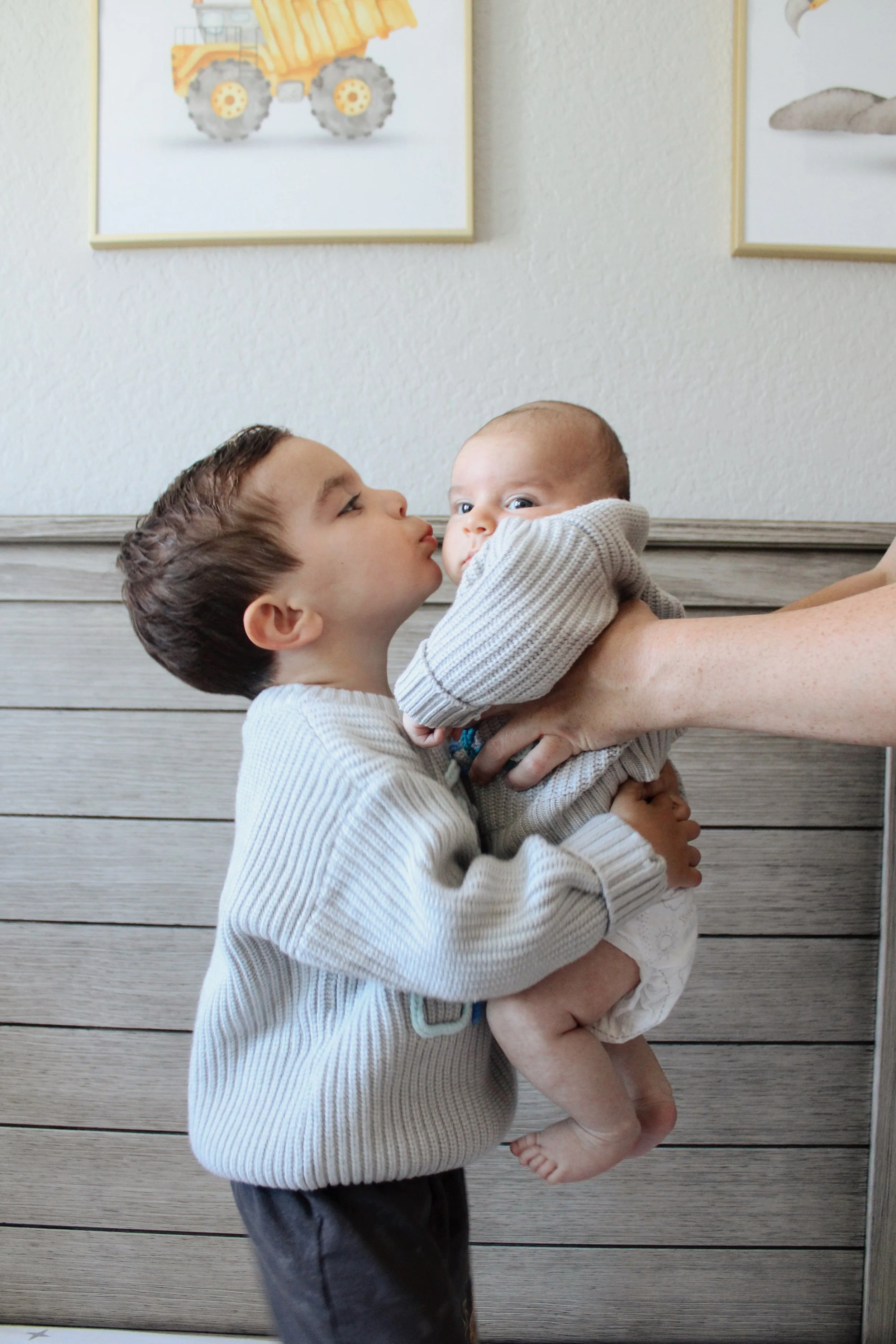 A young boy holding a baby, appearing to kiss or nuzzle the baby, in a room with framed pictures of construction vehicles on the wall.