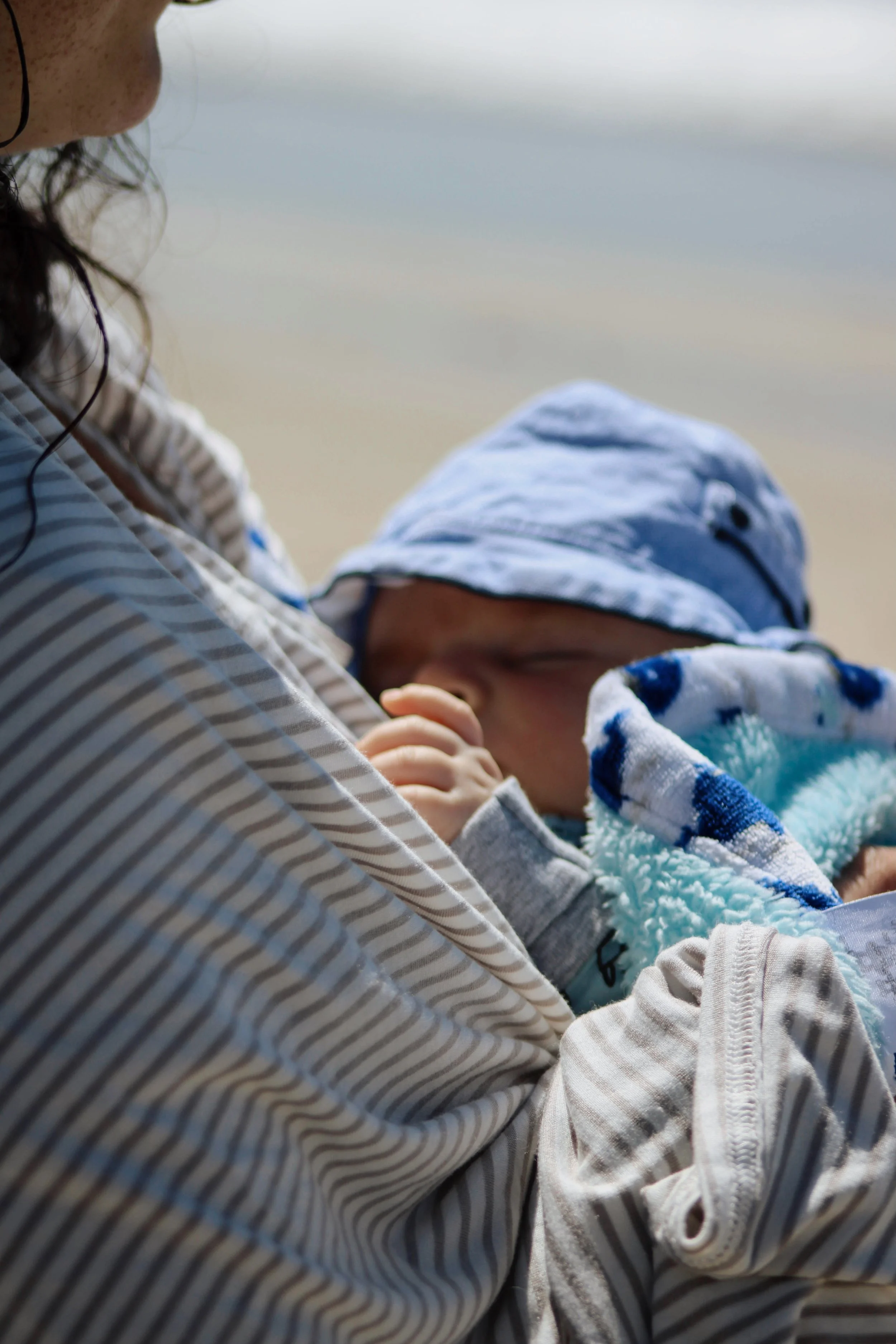 A woman holding a sleeping baby wrapped in a towel near the beach.