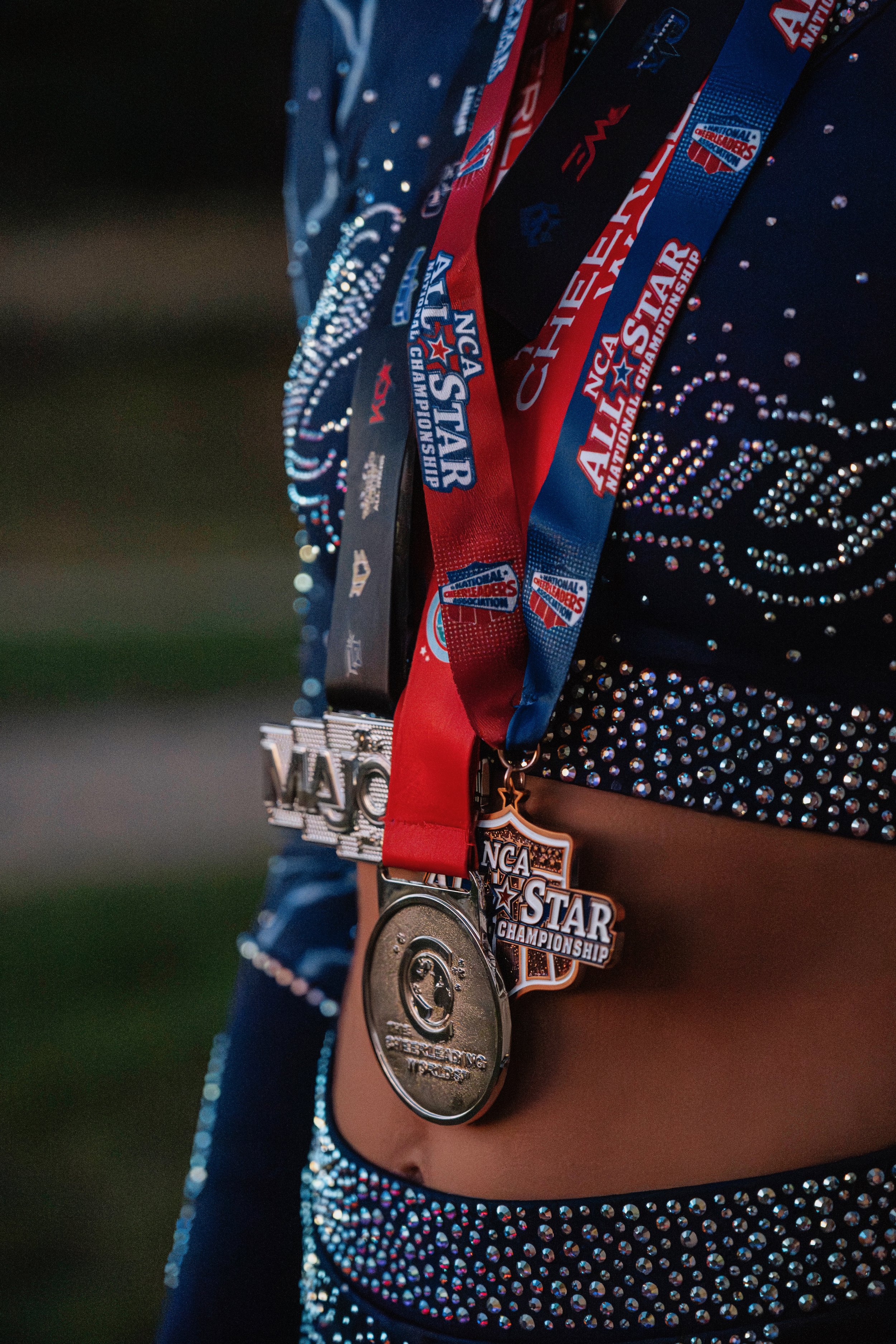 Close-up of a cheerleader's waist showing multiple medals, ribbons, and badges from the NCA All-Star Championship, with a dark background.