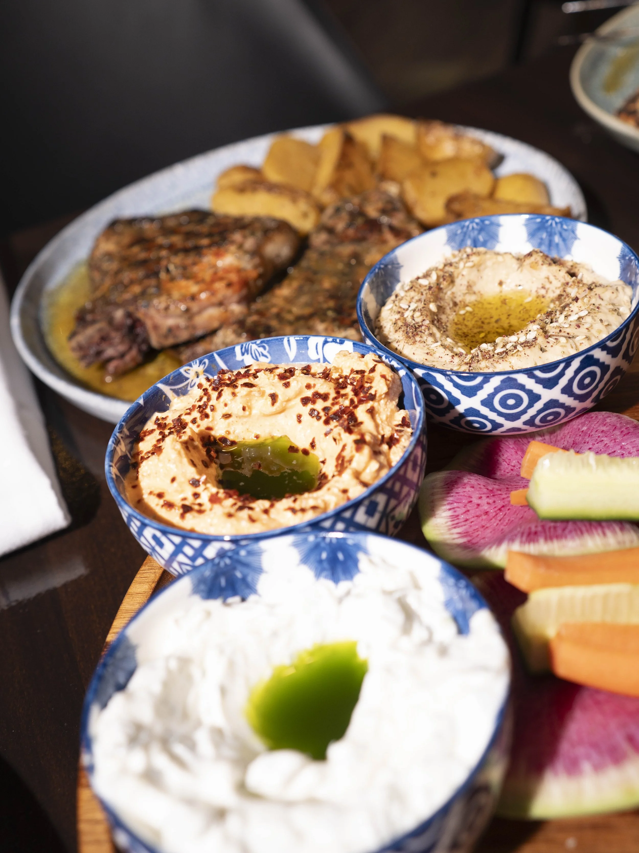 A platter of grilled meat with roasted potatoes and three bowls of dips, including hummus and other spreads, garnished with olive oil and herbs, along with sliced vegetables like watermelon radish, carrots, and cucumber.