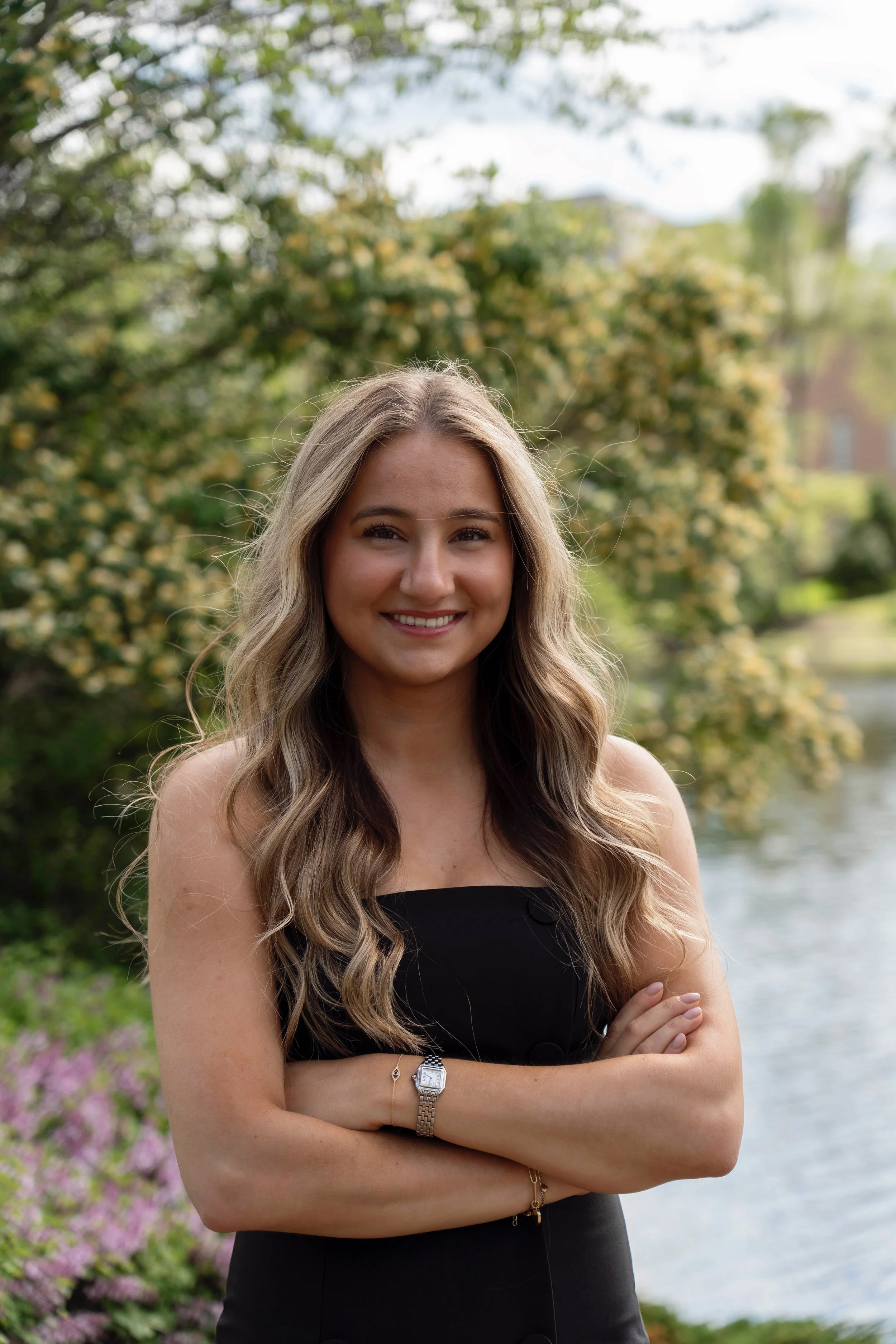 A smiling woman with long wavy blonde hair standing outdoors near a body of water, with green trees and flowering bushes in the background.