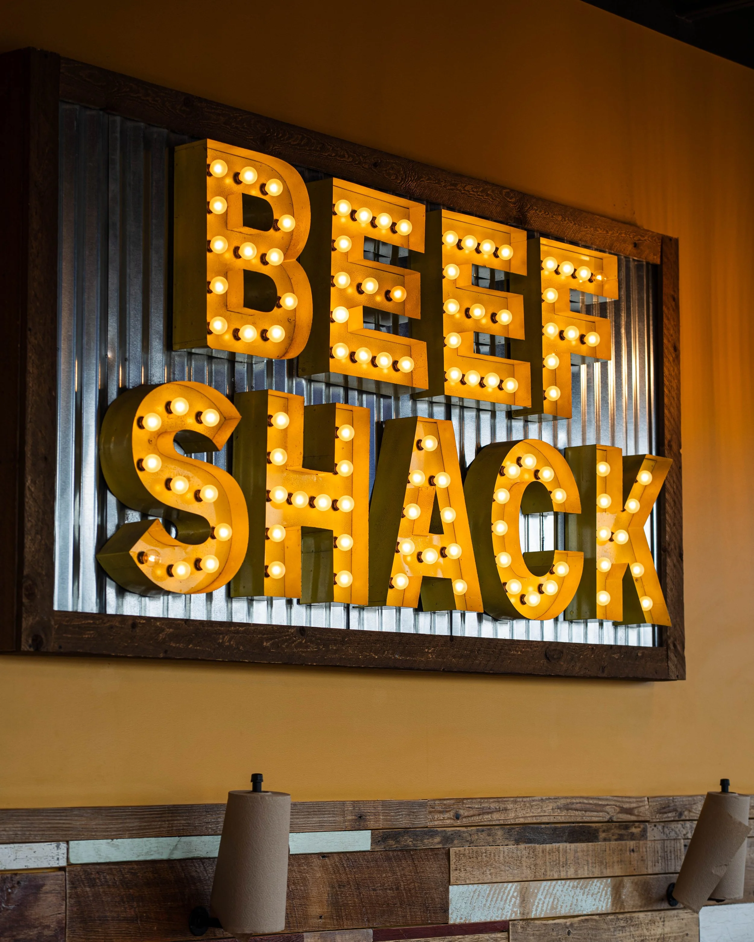 Decorative sign with the words 'Beer Shack' in large yellow letters with light bulbs, mounted on a wall with wood and metal paneling.