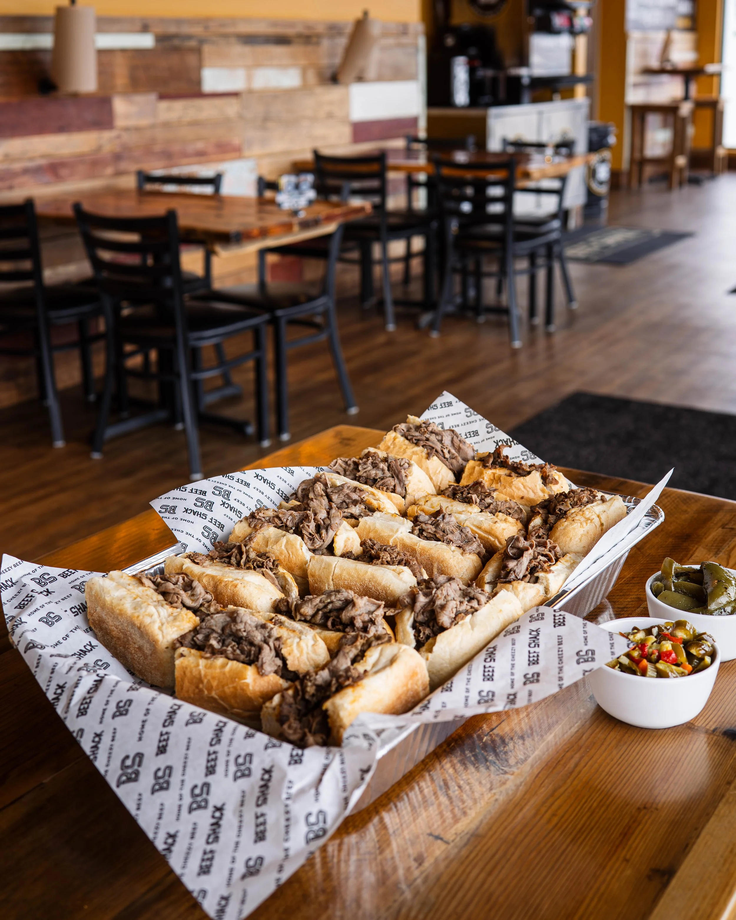 Beef sandwiches with shredded beef on hoagie rolls, served with pickles and a side of jalapeño peppers, on a wooden table in a casual restaurant.