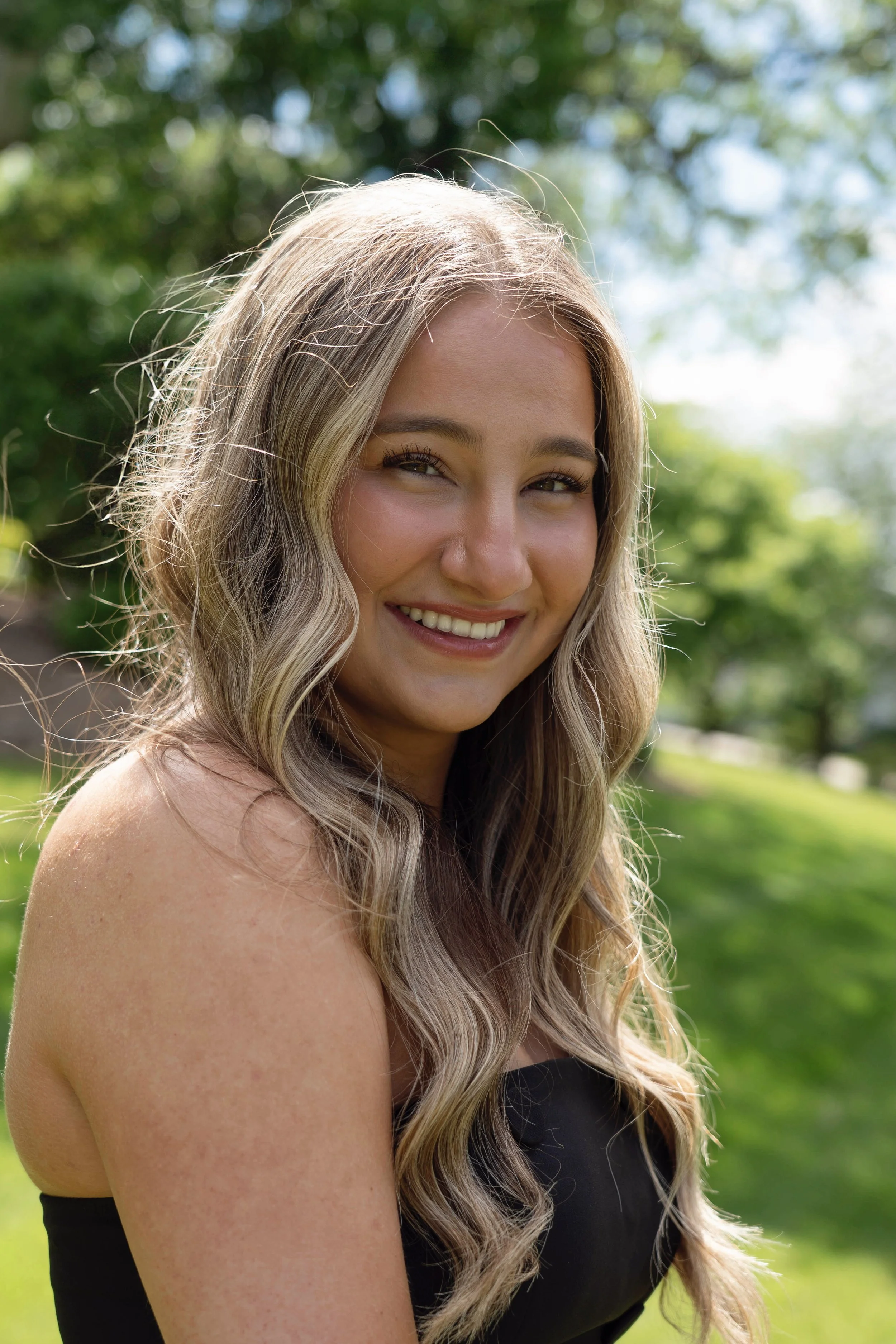 Close-up photo of a young woman with long, wavy blonde hair smiling outdoors, with greenery and trees in the background.