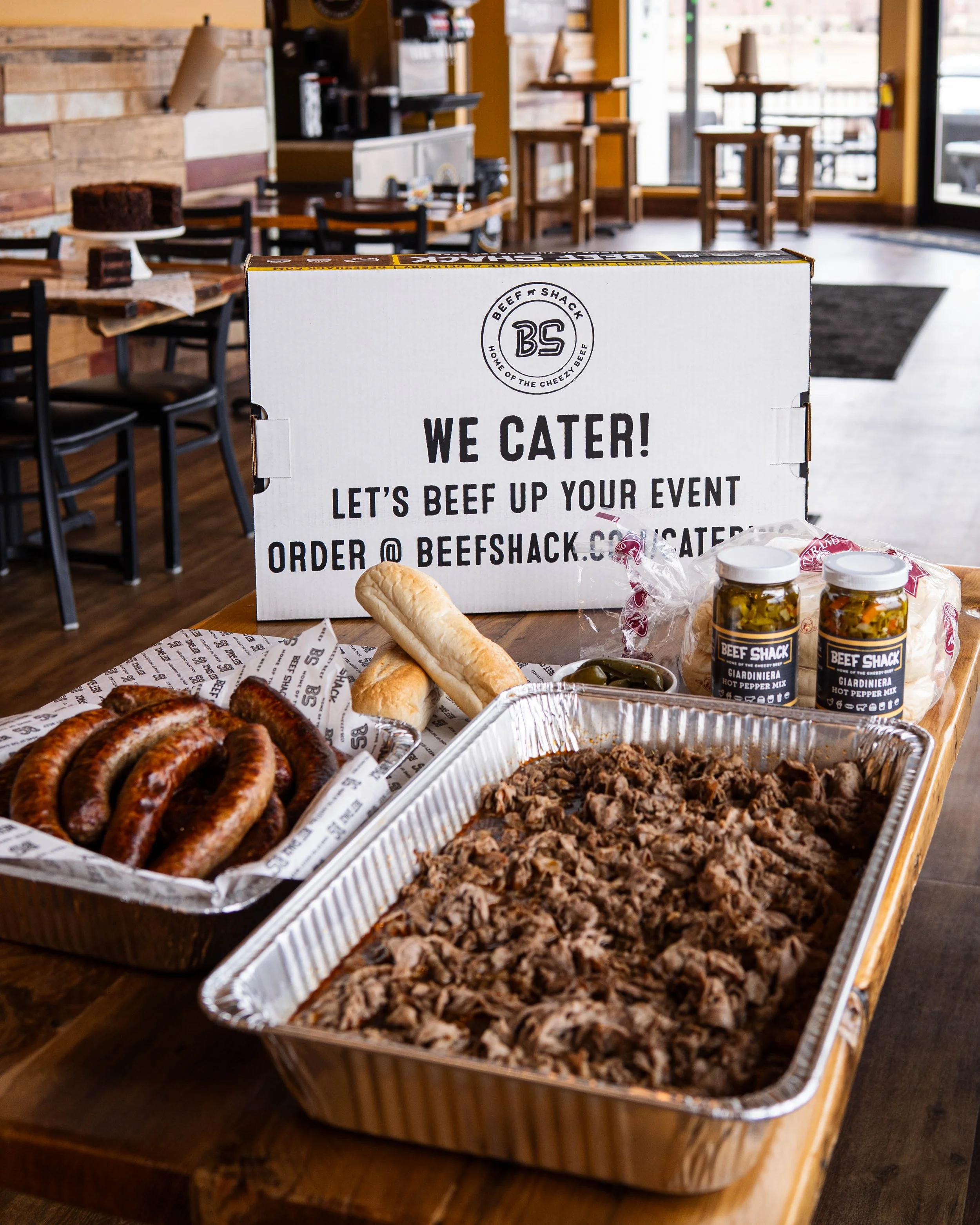 A table with large aluminum trays of cooked beef and sausages, two jars of hot pepper mix, and bread rolls, set in a restaurant with wooden decor and a sign reading 'We Cater! Let's beef up your event order @ beefshack.com'.