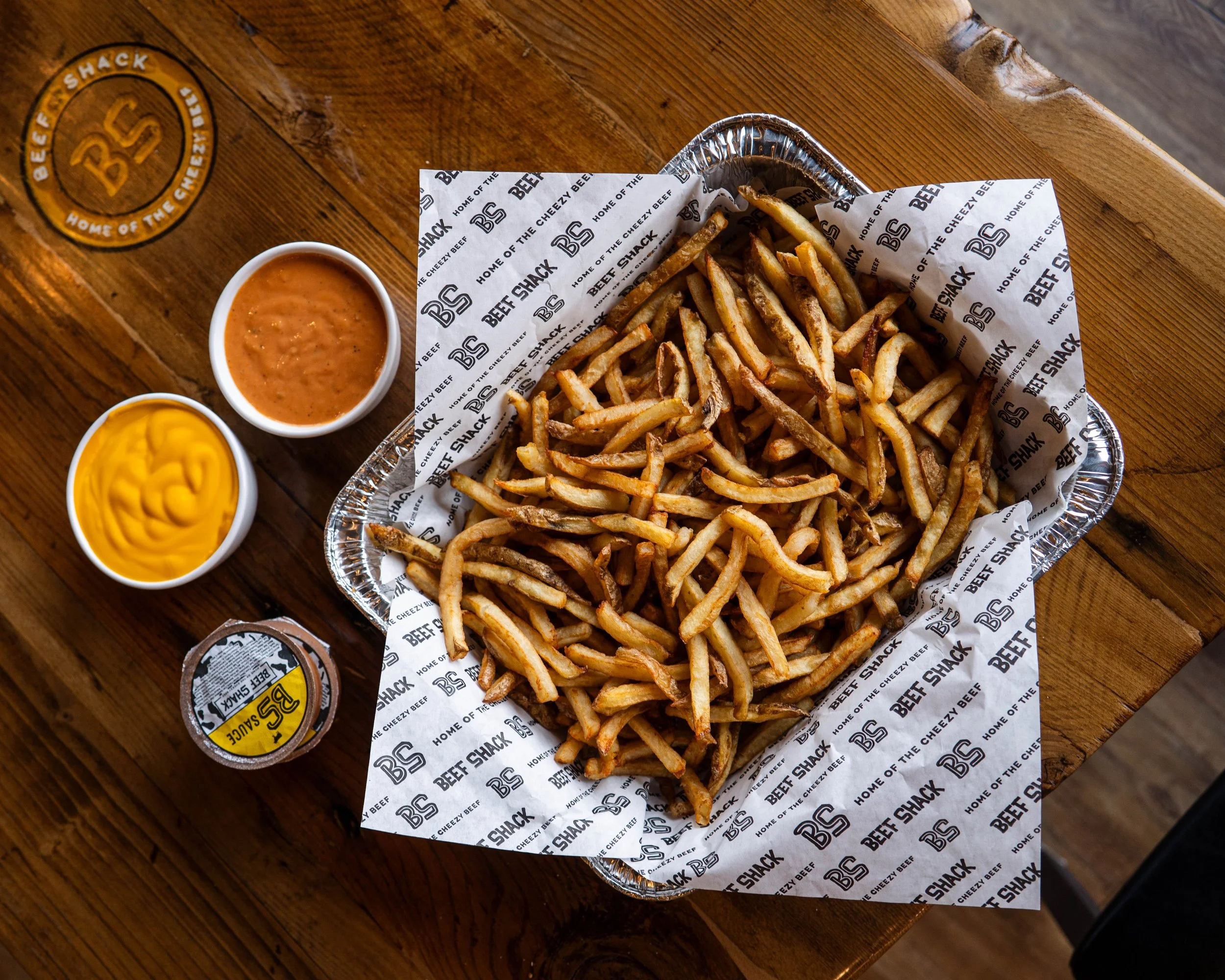 Tray of French fries with two dipping sauces, one yellow mustard and one reddish barbecue, on a wooden table with a container of barbecue sauce and a coin-shaped logo that reads 'BEF SHACK'.