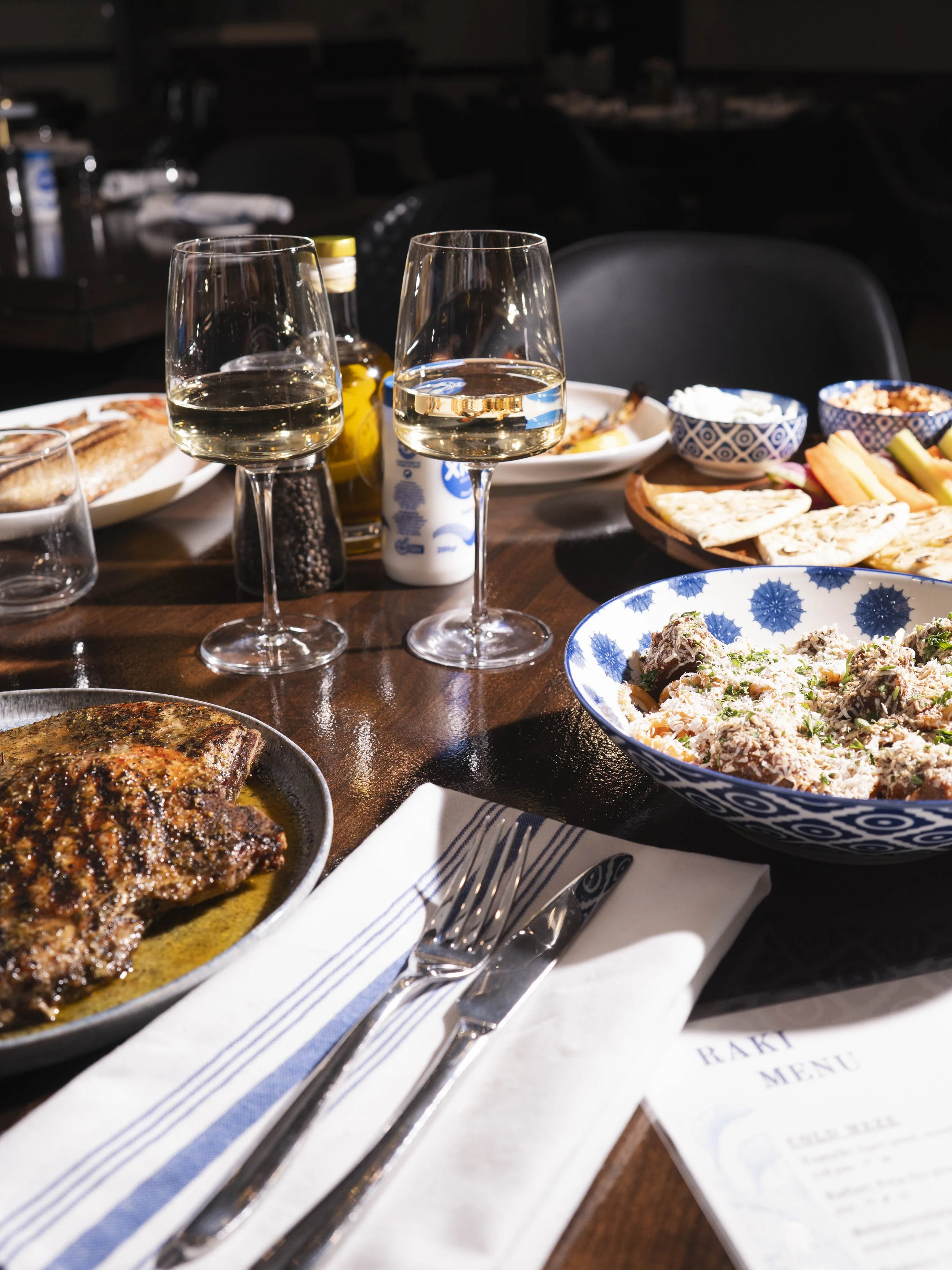 A table set with two glasses of white wine, a plate of cooked meat, various side dishes in bowls, and a menu, with a dark background.