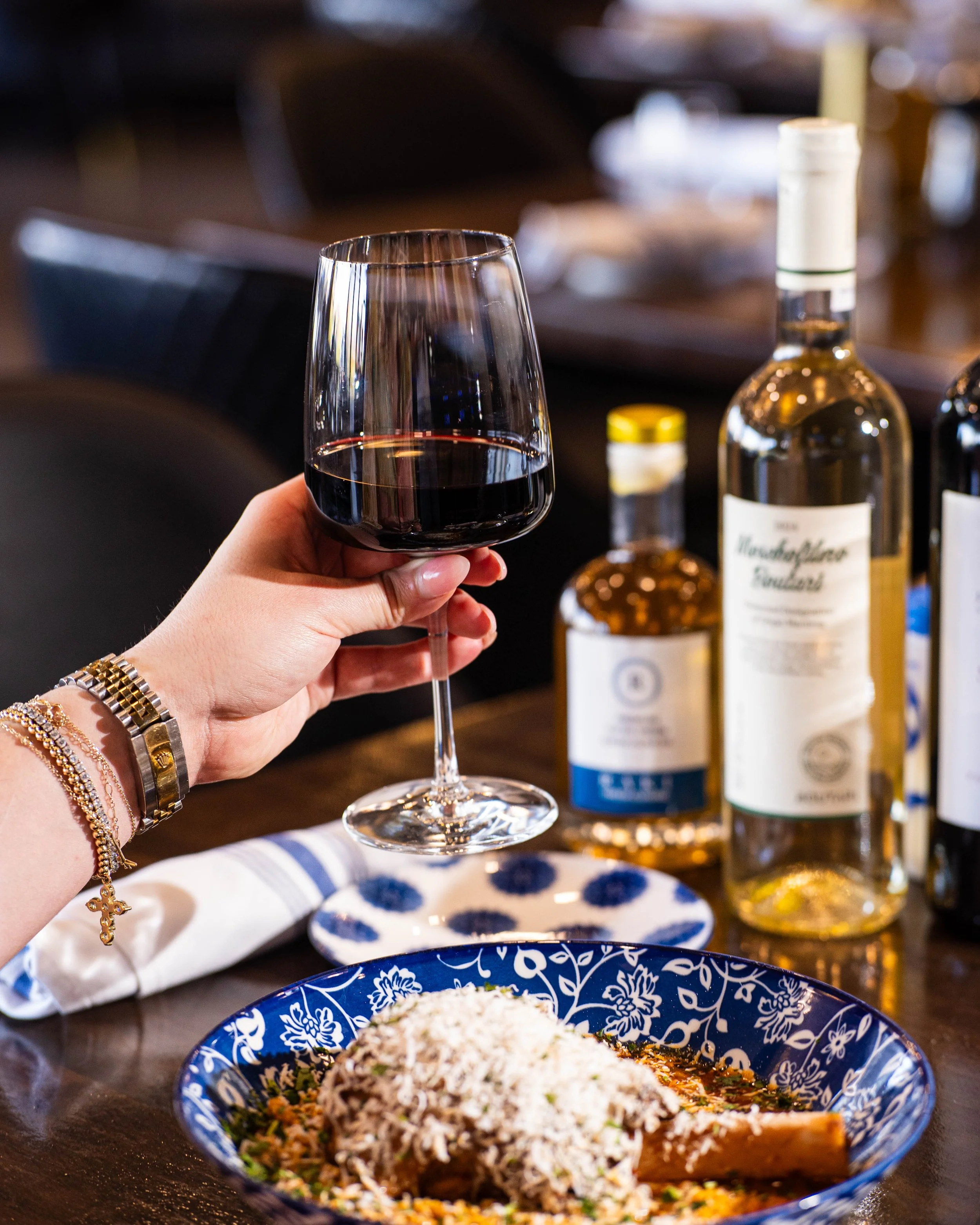 A person holding a glass of red wine next to a plate of food, with bottles of alcohol on the table.