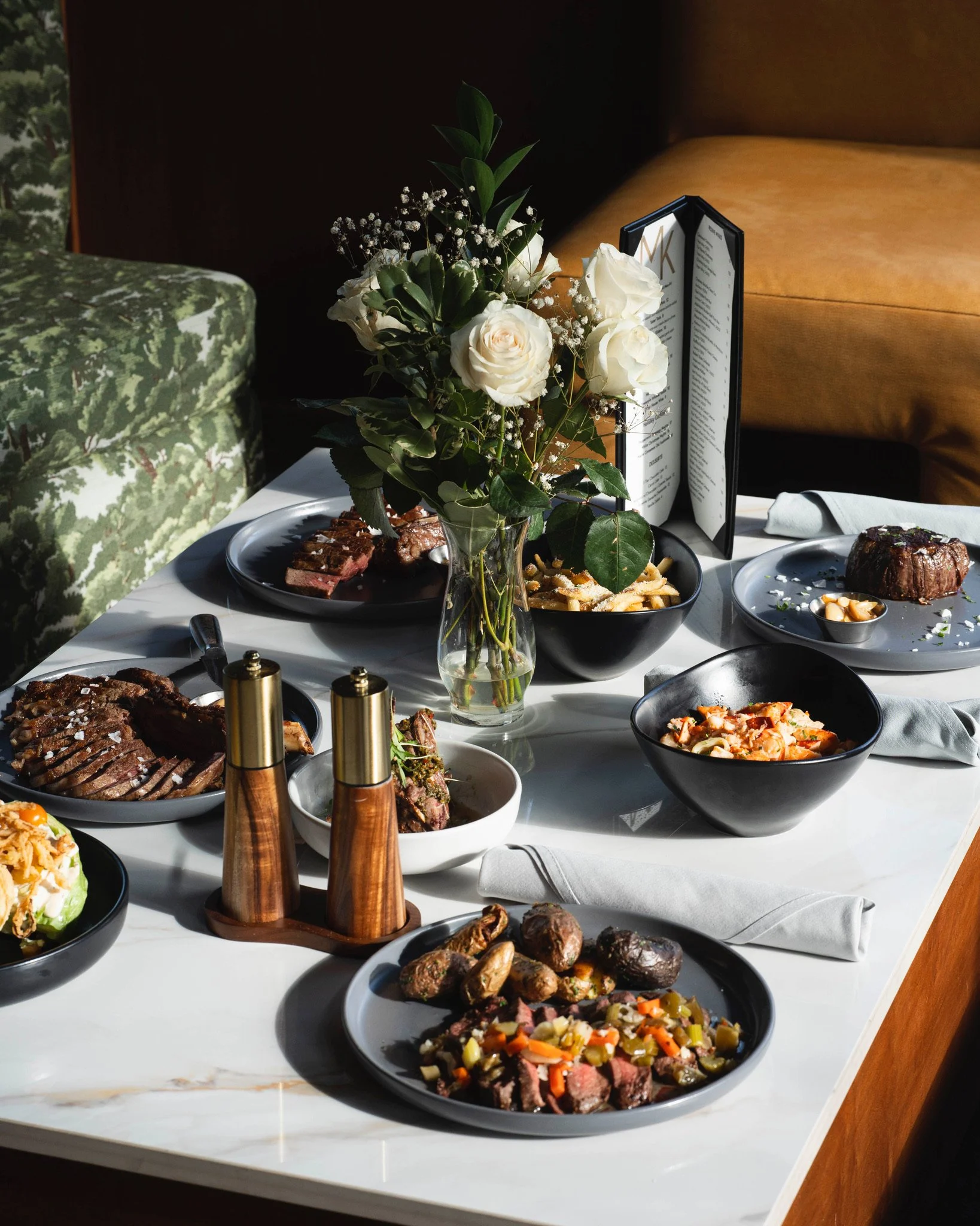 A table set for a meal with various dishes, a vase of white roses, a menu, and black bowls with food, on a white marble table with gray napkins.