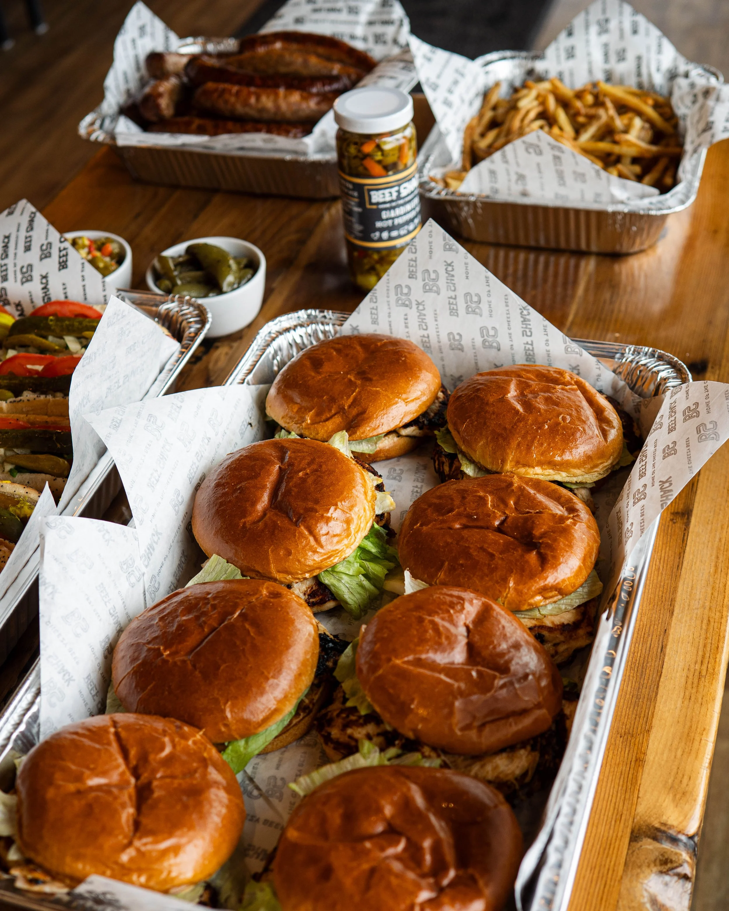 Several beef sliders with lettuce in a foil tray at a rustic wooden table, with sides of fries, pickles, and other condiments and sauces in the background.