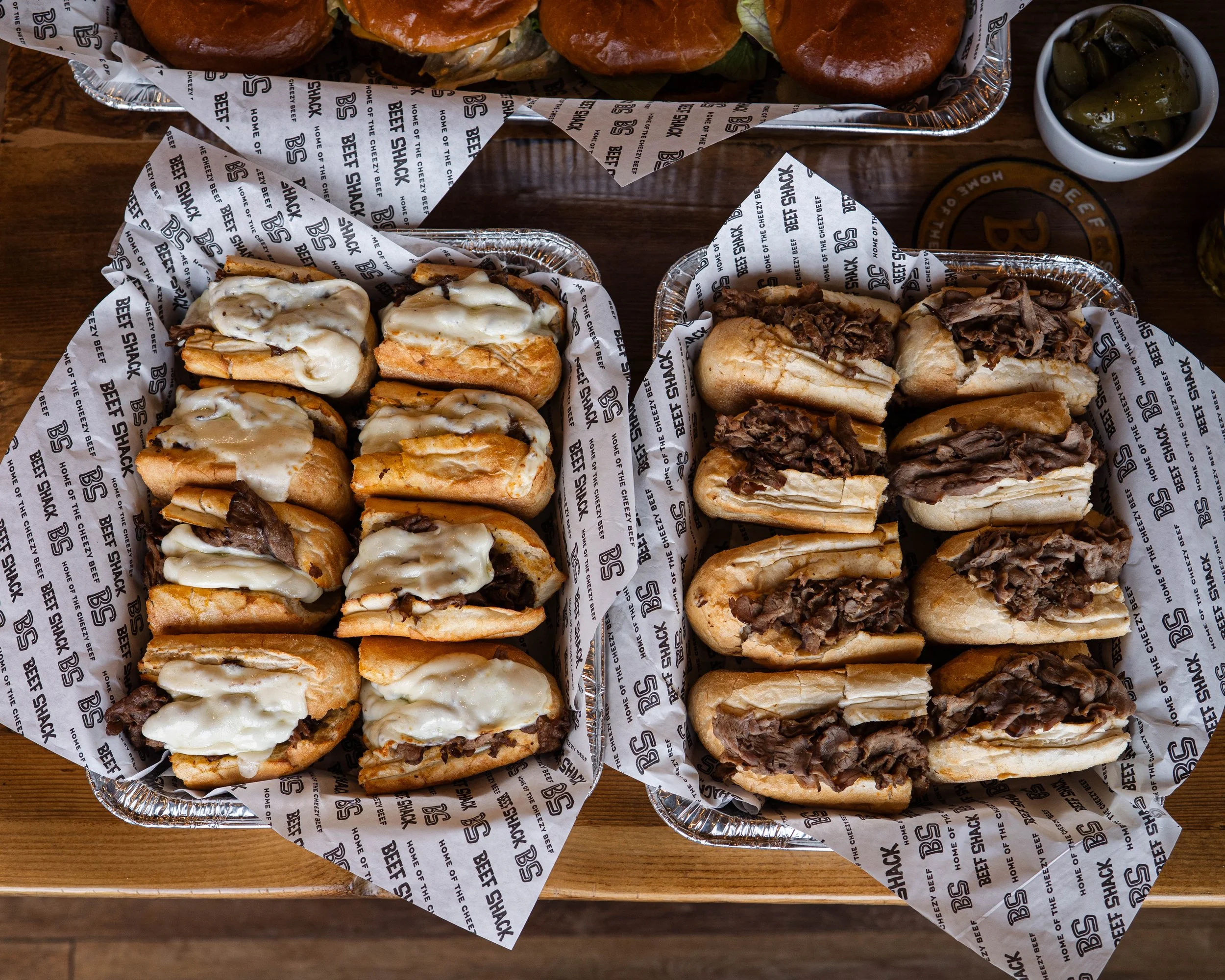 Two trays of sandwiches on a wooden table, with pickles in the background. The tray on the left has sandwiches with melted cheese and beef, while the tray on the right has sandwiches filled with shredded beef.