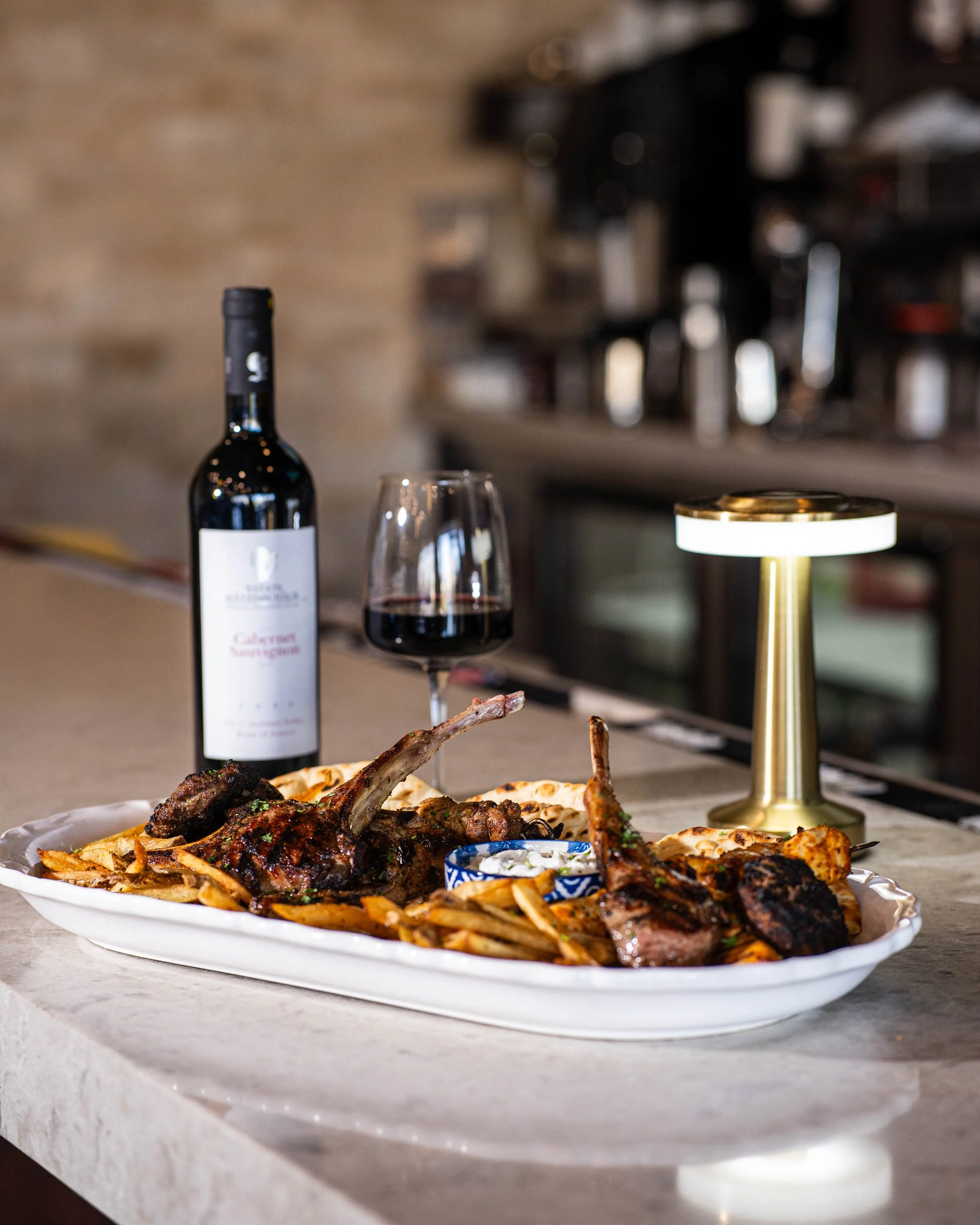 A dinner plate with lamb chops and fries, a glass of red wine, a bottle of red wine, and a small bowl of sauce on a marble countertop, with a small gold lamp and a blurred kitchen background.