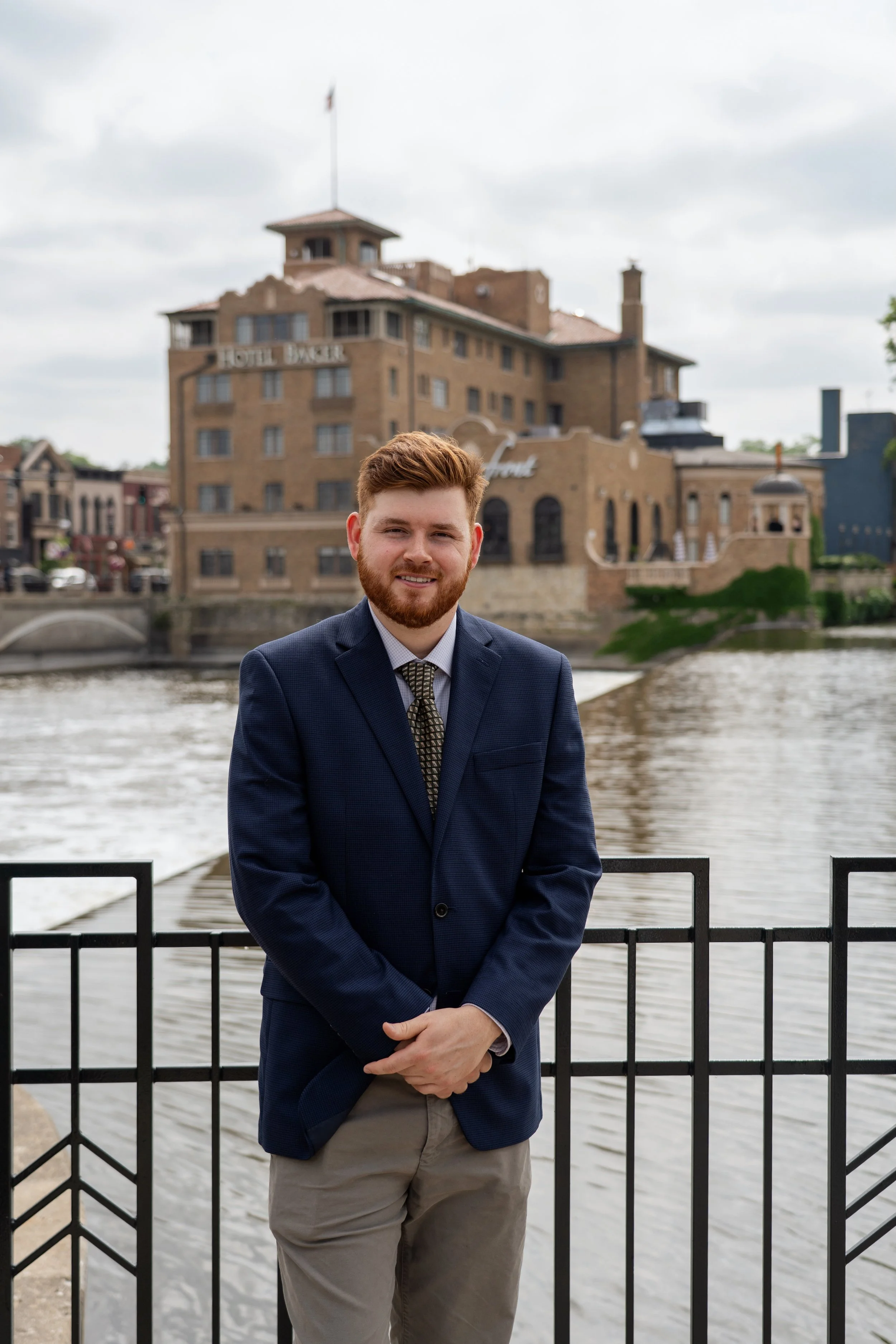 A man in a navy blazer and khaki pants standing outdoors by a river, with a historic brick building labeled 'Hotel Baker' in the background.