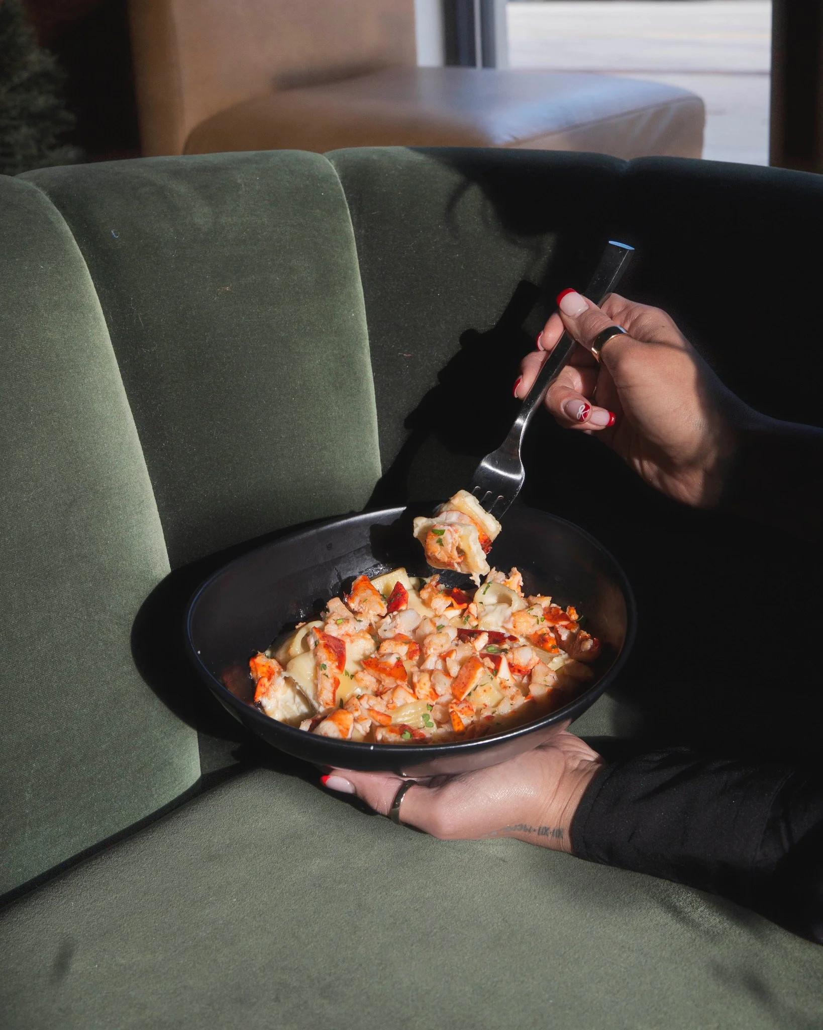 A person holding a fork and a black bowl of cooked seafood pasta on a green sofa.