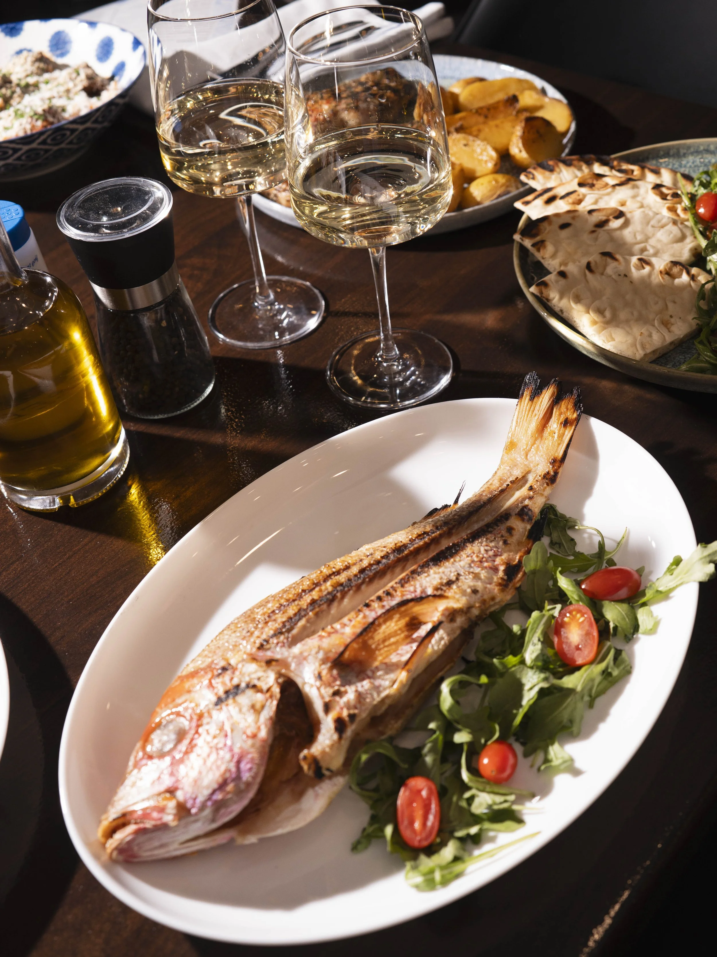 Grilled fish served on a white plate, garnished with greens and cherry tomatoes, accompanied by glasses of white wine, with various side dishes including potatoes, flatbread, salad, and rice on a dark wooden table.