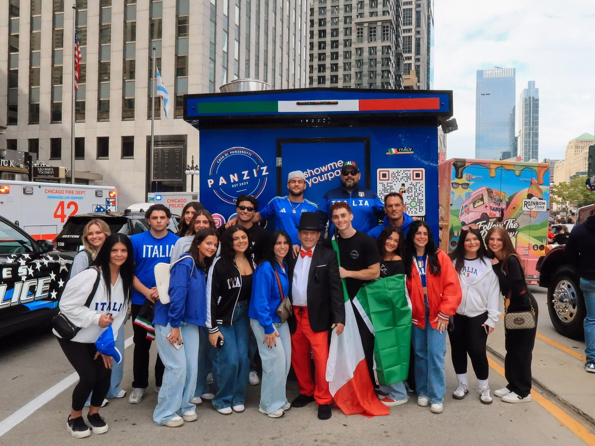Group of people gathered in front of a blue food truck with Italian flags and police cars nearby, in an urban city setting.