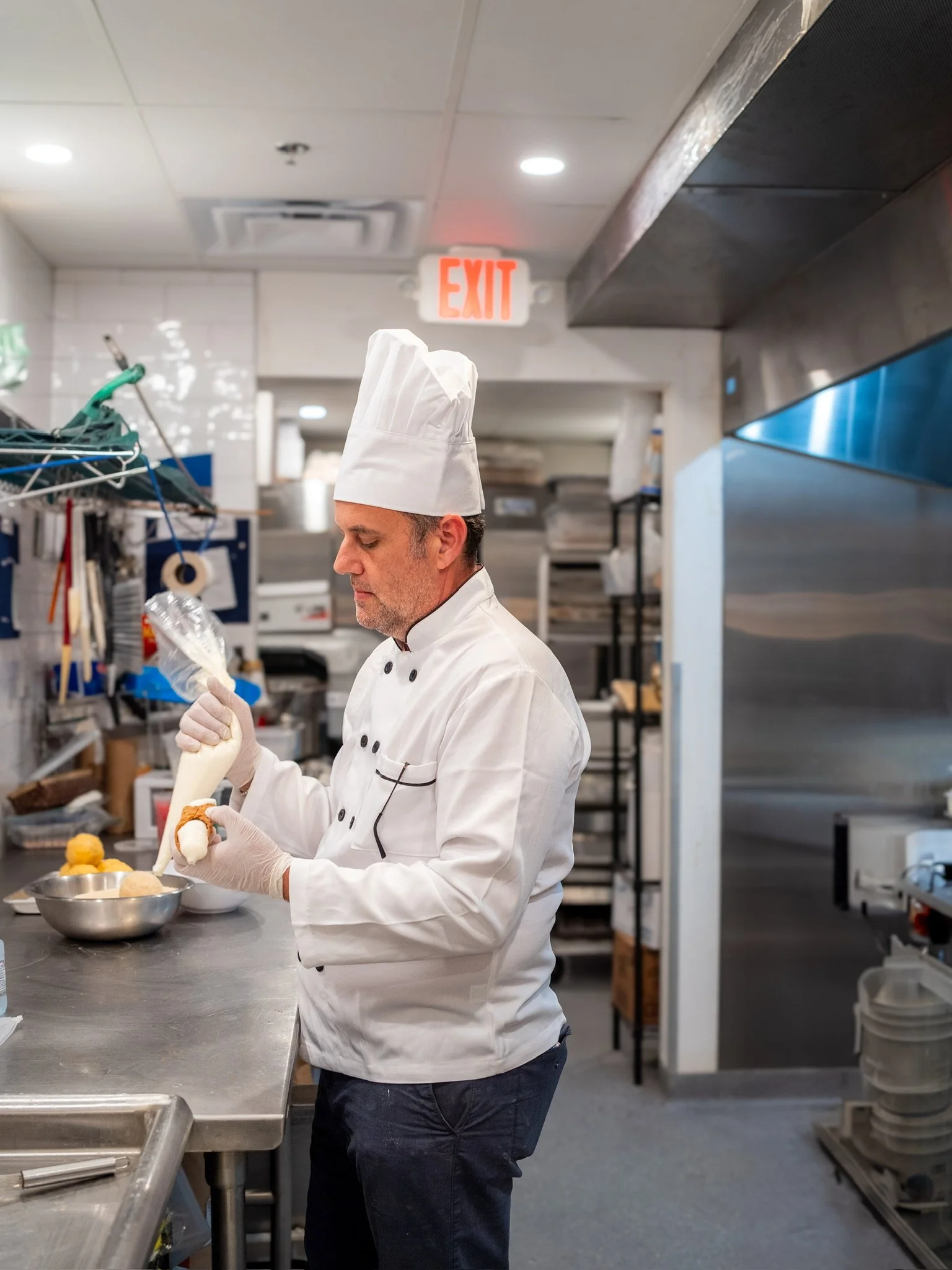 Chef in white uniform and hat squeezing cream from a pastry bag into a dessert in a commercial kitchen.