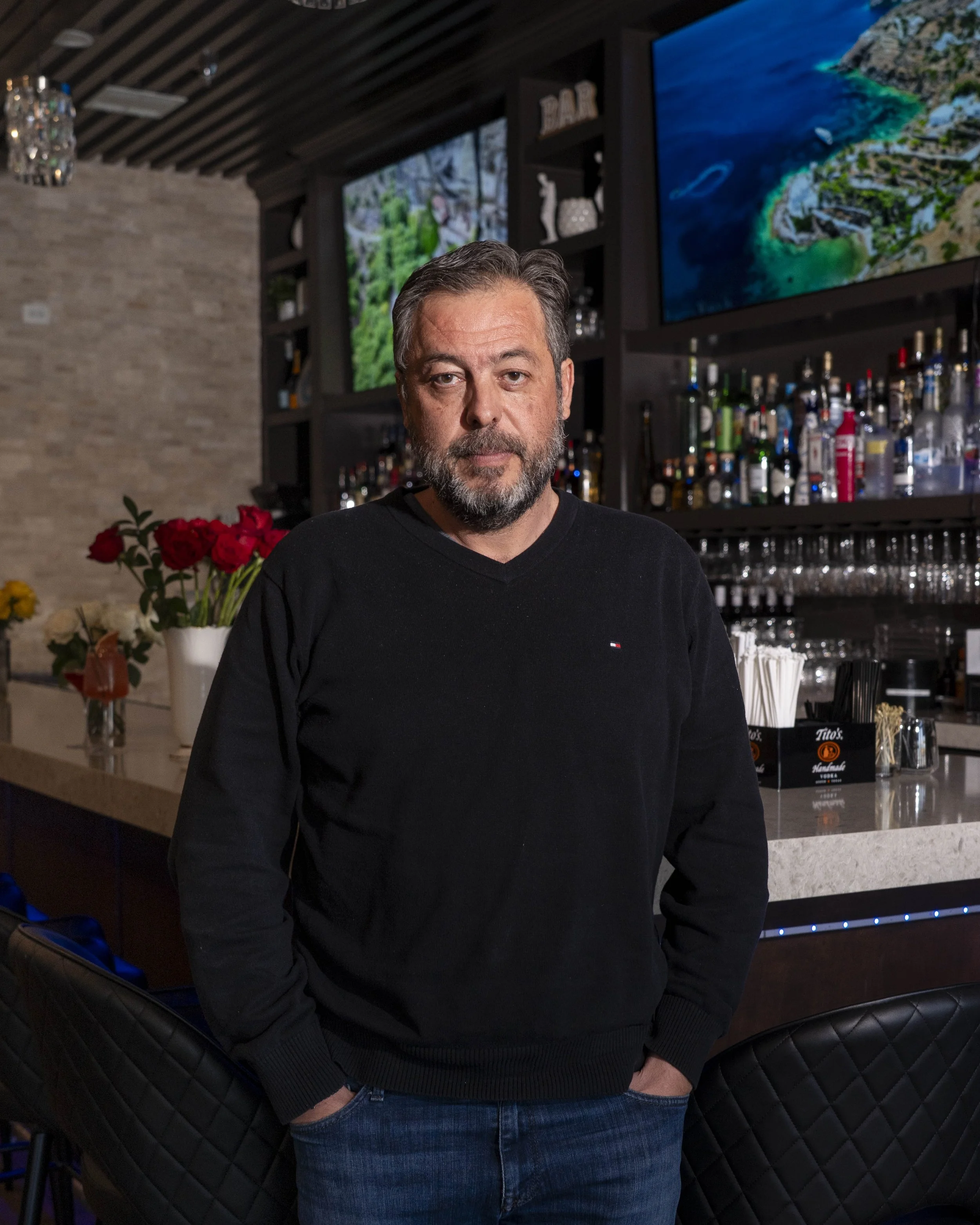 Man with gray hair and beard standing in a bar with liquor bottles behind him and TV screens above, flowers on the bar counter.