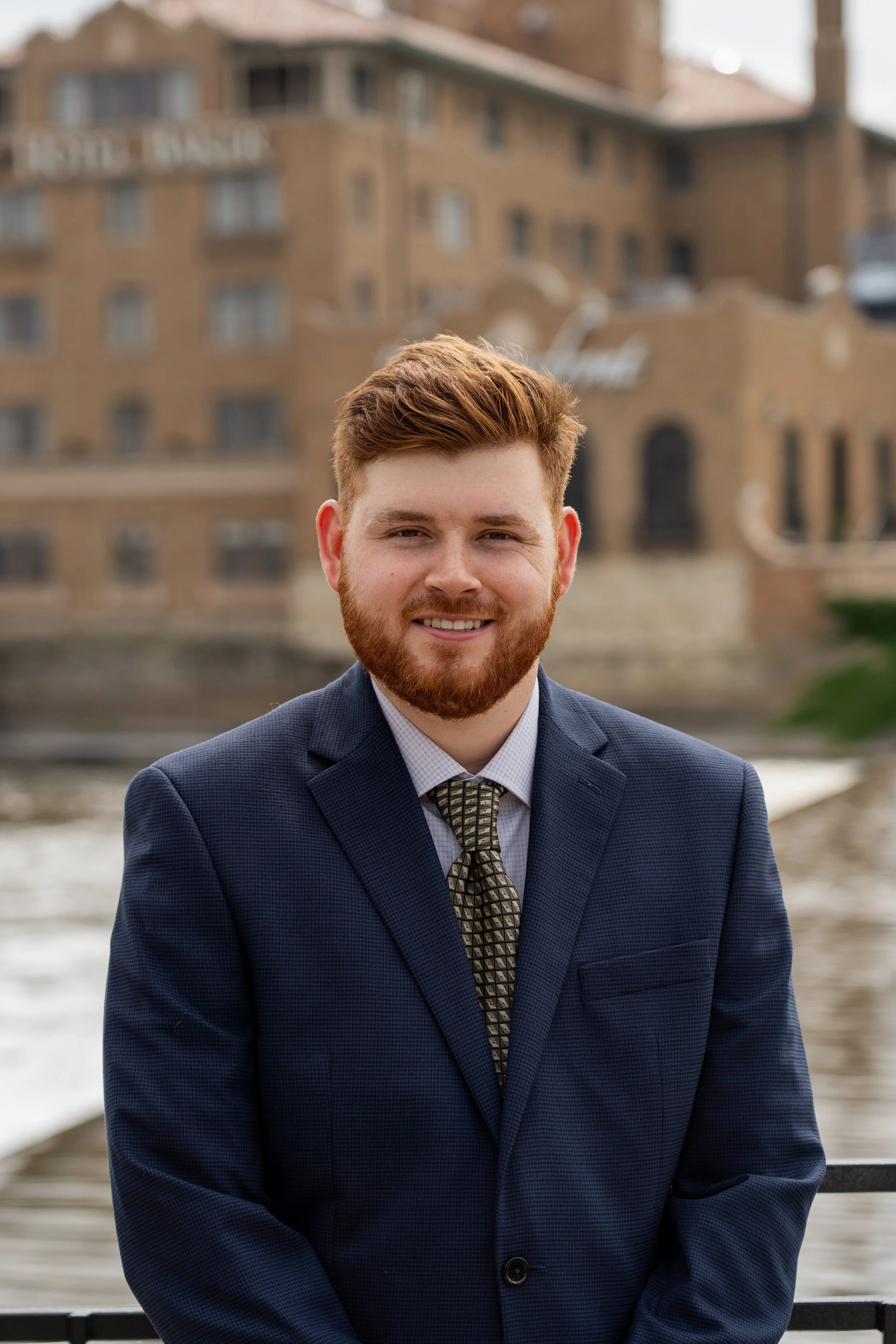 A smiling man with red hair and beard in a navy suit and patterned tie, standing outdoors near a river with a historic building in the background.