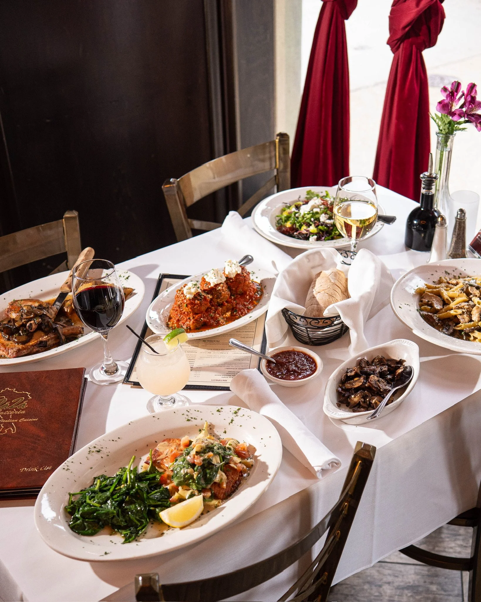 A table set with various plates of food, wine glasses, a cocktail, bread, and condiments in a restaurant.