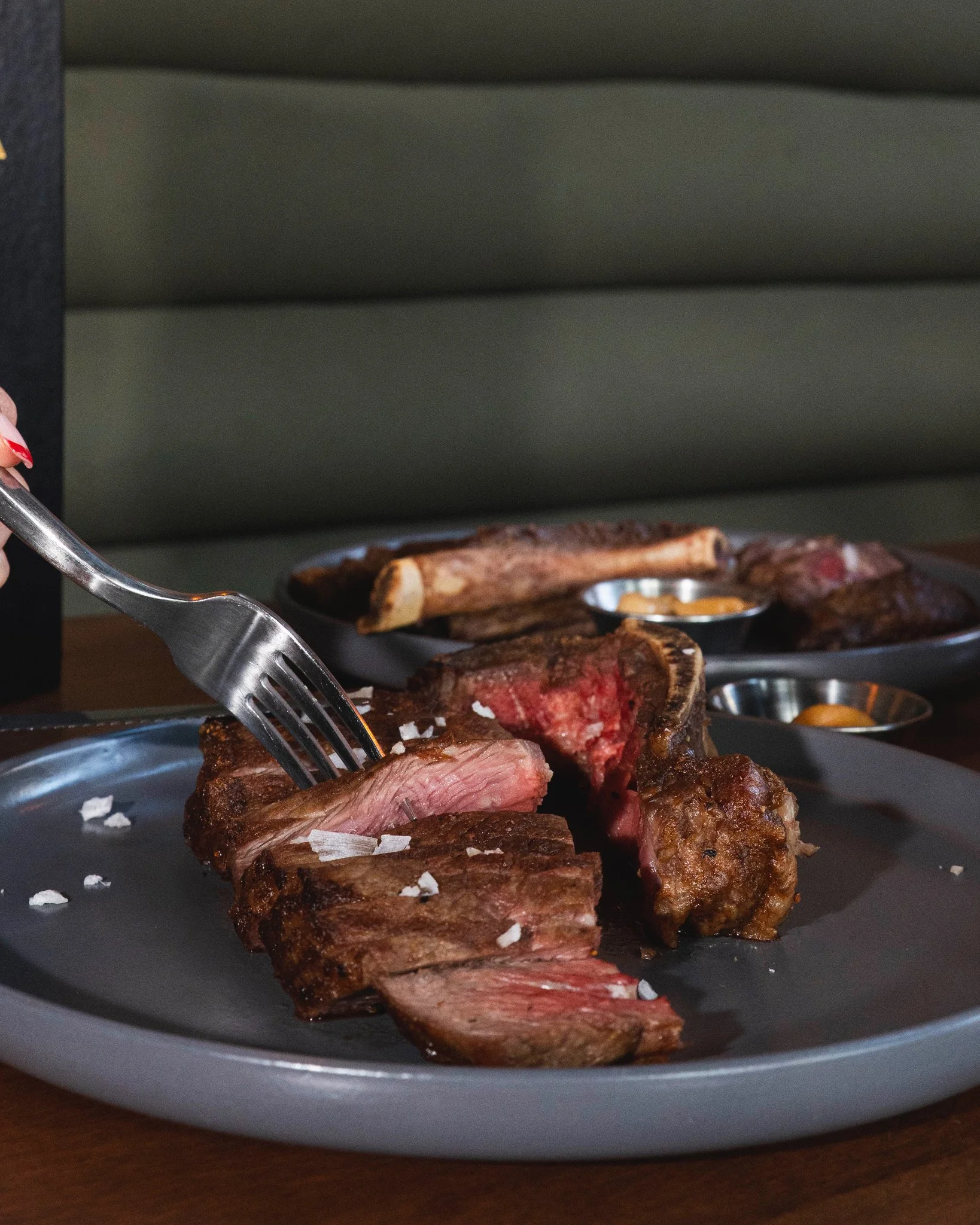 A plate with sliced, cooked steak and salt flakes, with additional meat cuts and dipping sauces in the background.