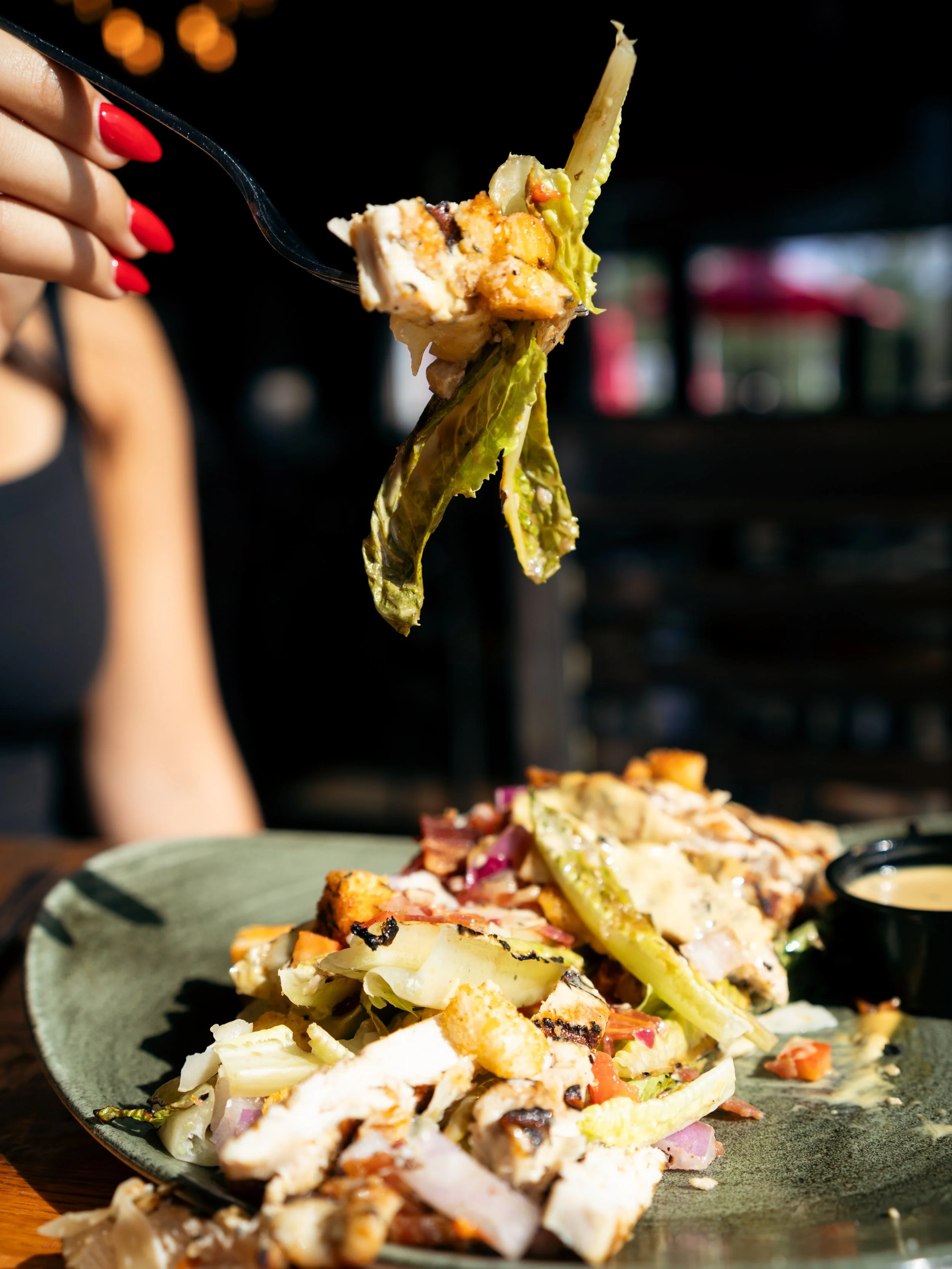 A fork holding a bite of salad with lettuce, grilled chicken, and vegetables above a plate of salad with grilled chicken and vegetables, with a small container of dressing on the side.