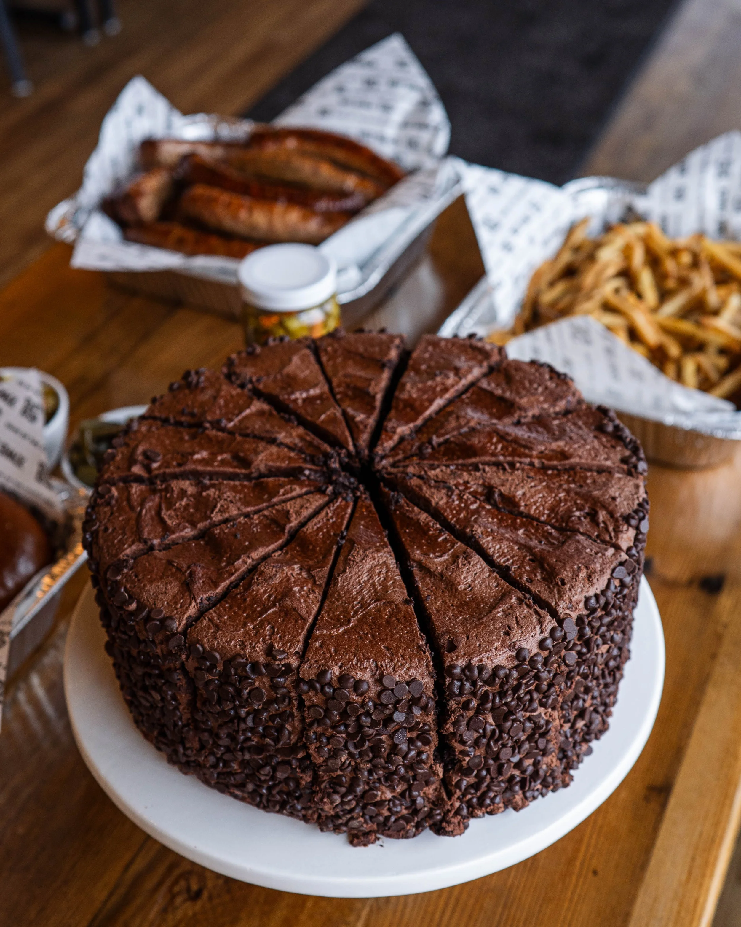 A large round chocolate cake with chocolate chips on the sides, sliced into pieces, on a white plate.