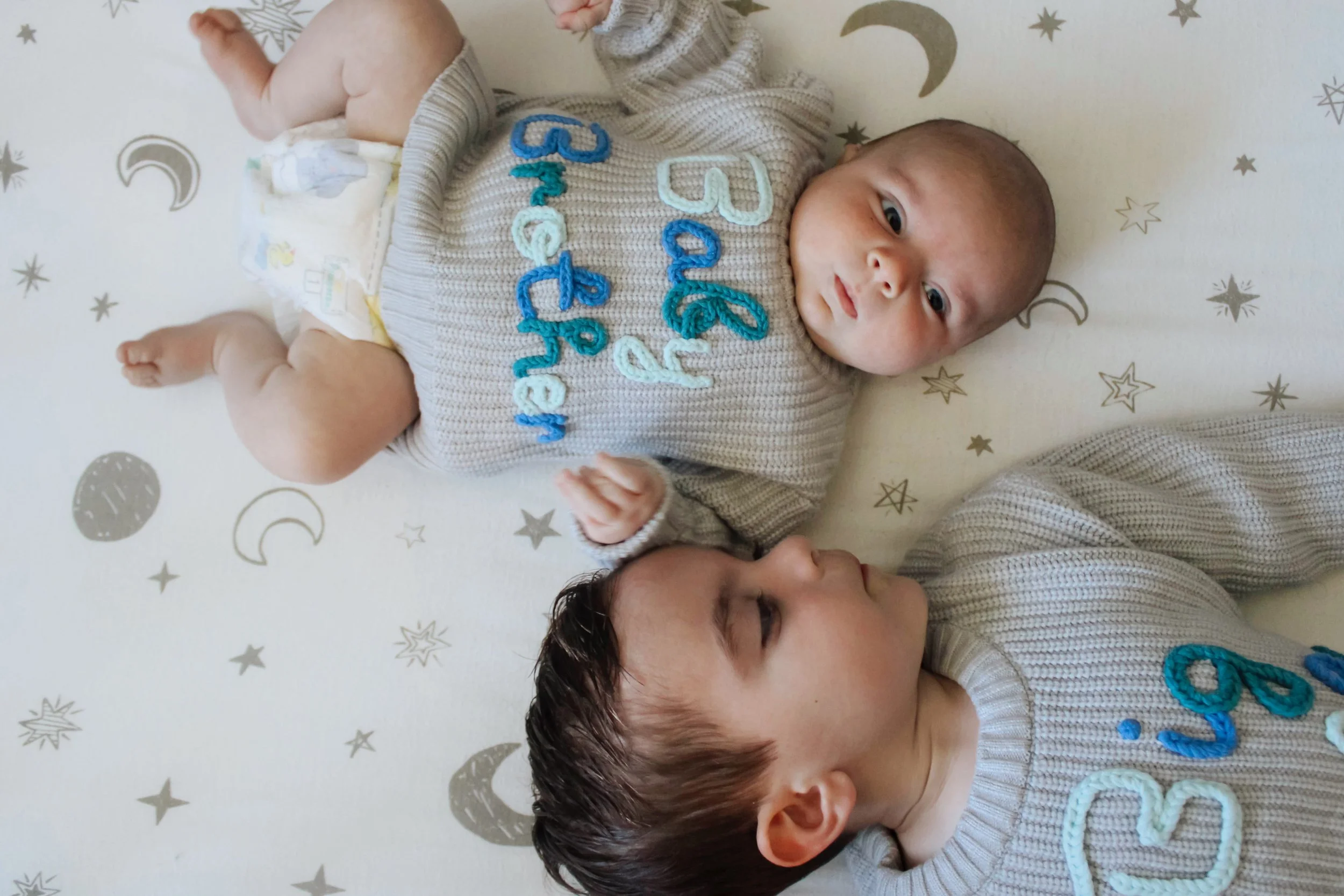 A baby and a young child lying on a blanket with moon and star patterns, both wearing matching knitted gray shirts with blue and white embroidered text.