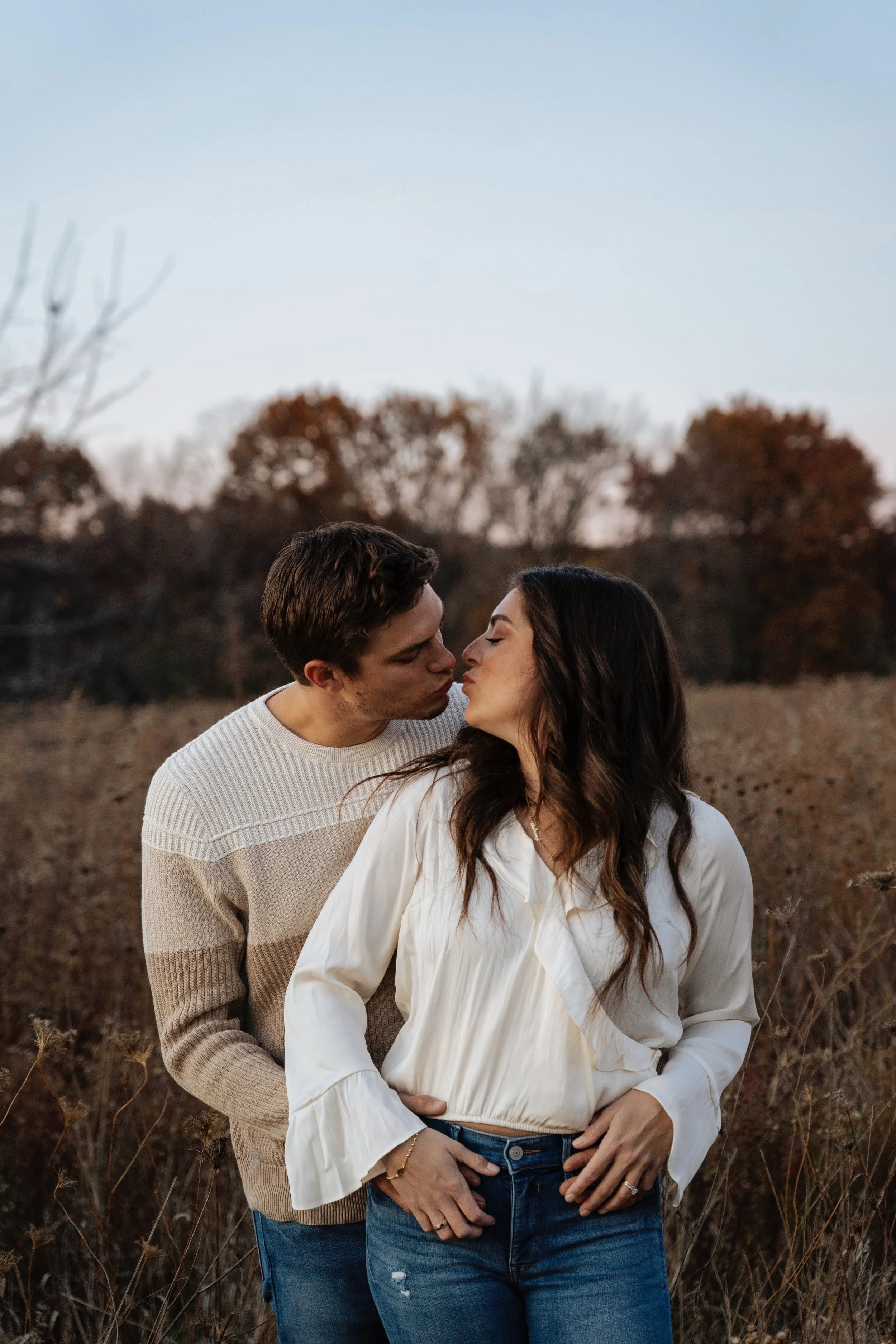 A couple sharing a kiss outdoors during autumn, standing in a field with trees and a sunset.