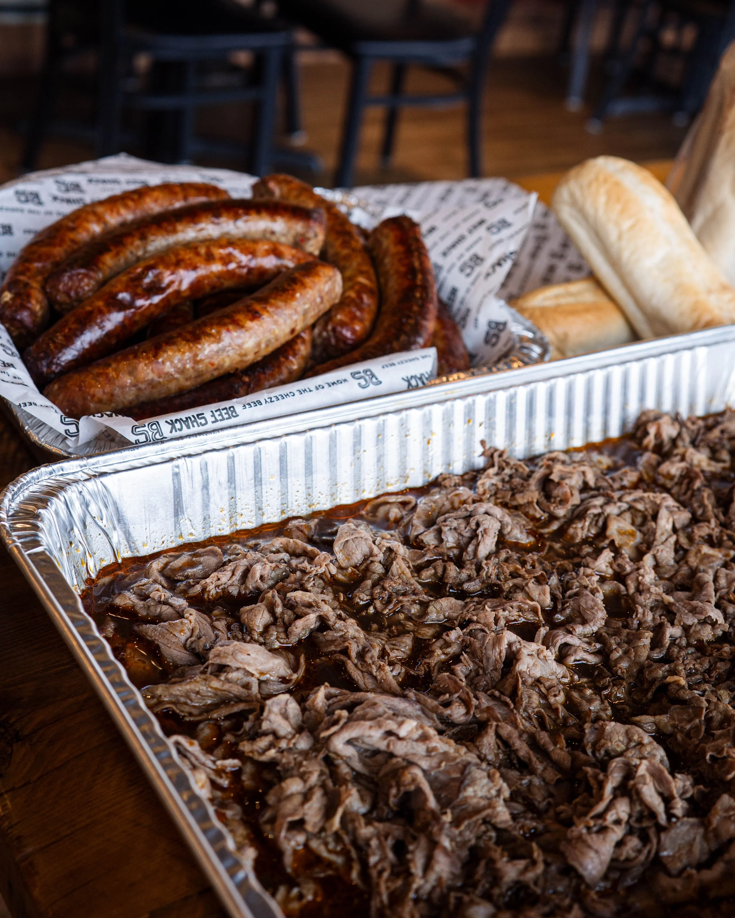 Barbecue platter with sliced beef and grilled sausages, accompanied by bread rolls.