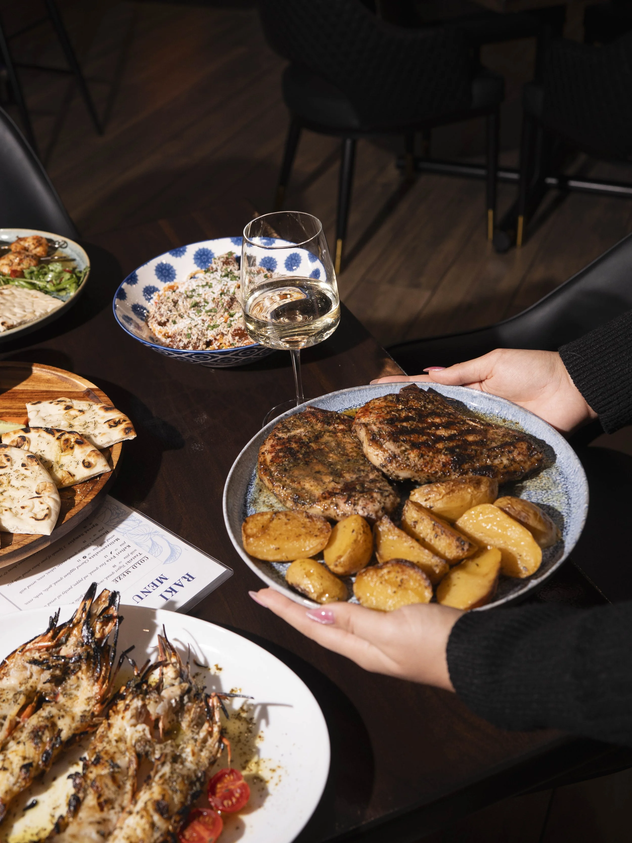 Person holding a plate with grilled steak and roasted potatoes at a dinner table with various dishes and a glass of white wine.