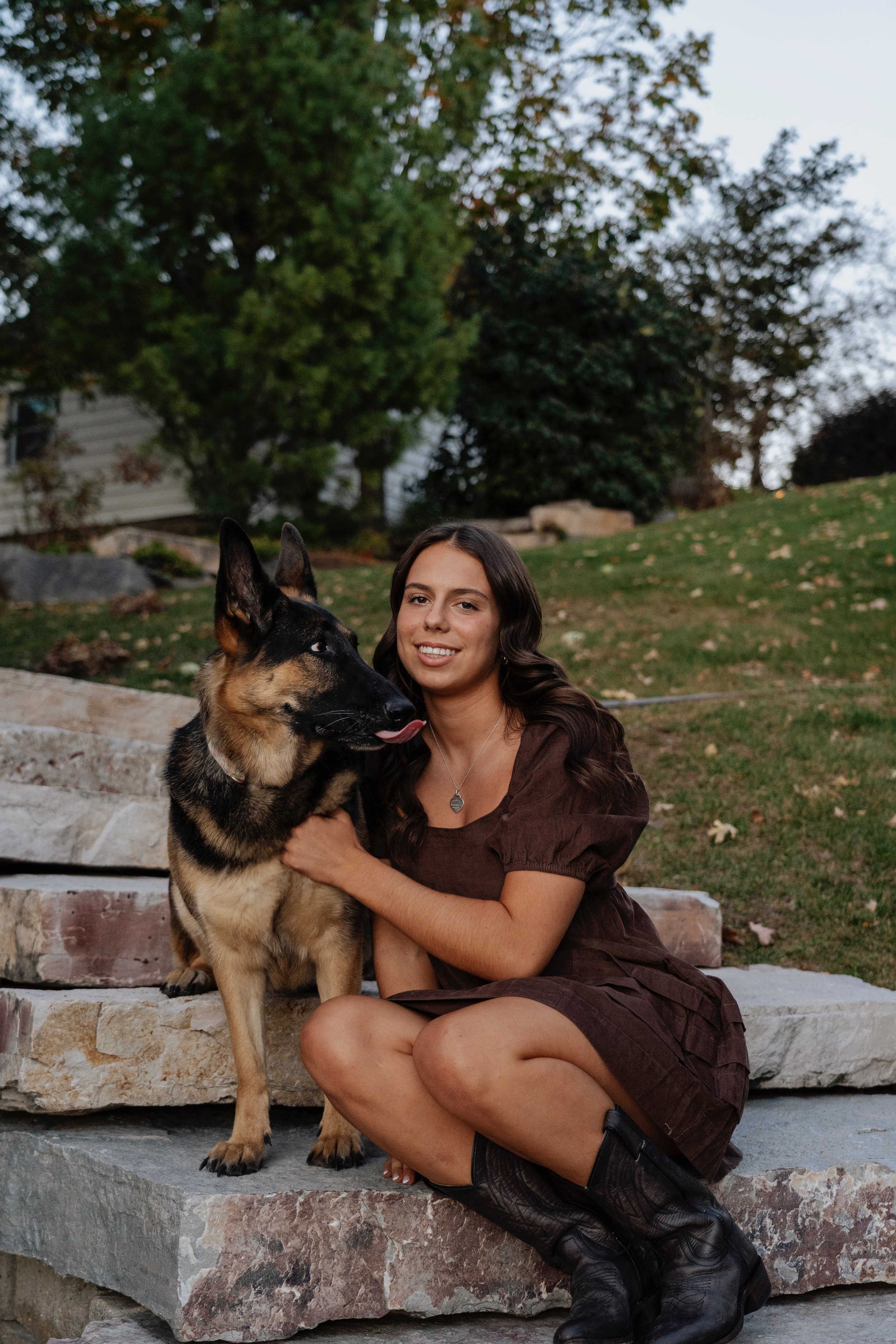 A woman with long dark hair, wearing a brown dress and black boots, sitting on stone steps outdoors, hugging a German Shepherd dog. The background shows trees and a grassy yard.