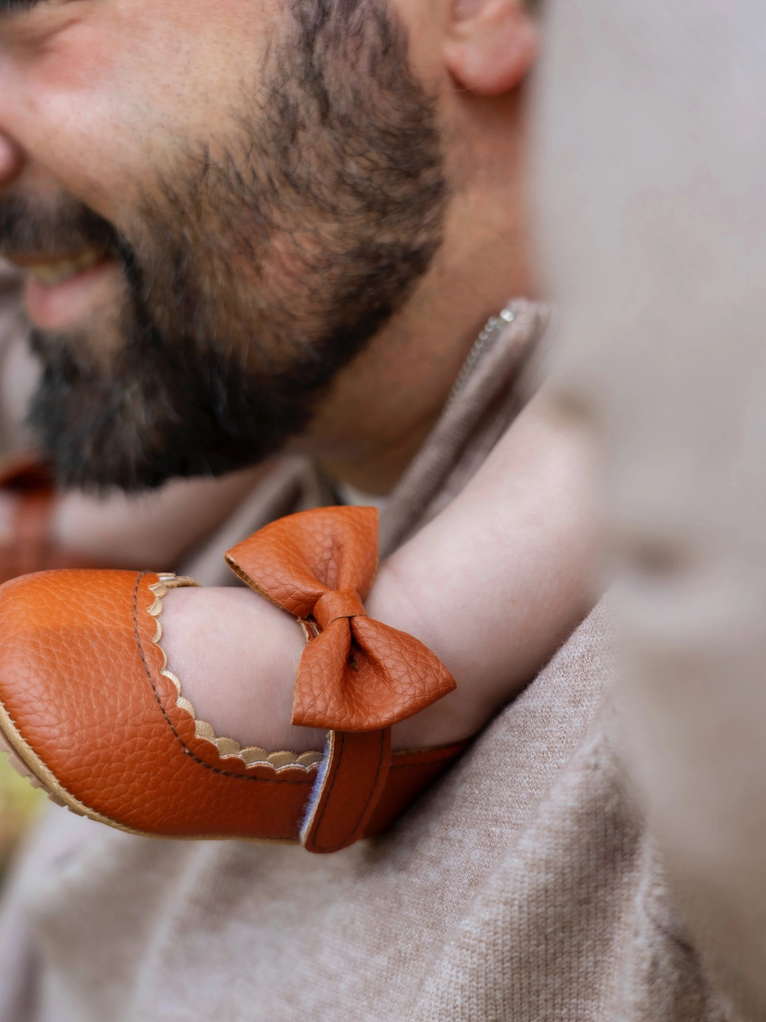 Close-up of a smiling man with a beard wearing a beige coat, with a young girl in a beige coat and orange leather shoes with a bow, holding hands close to his face.