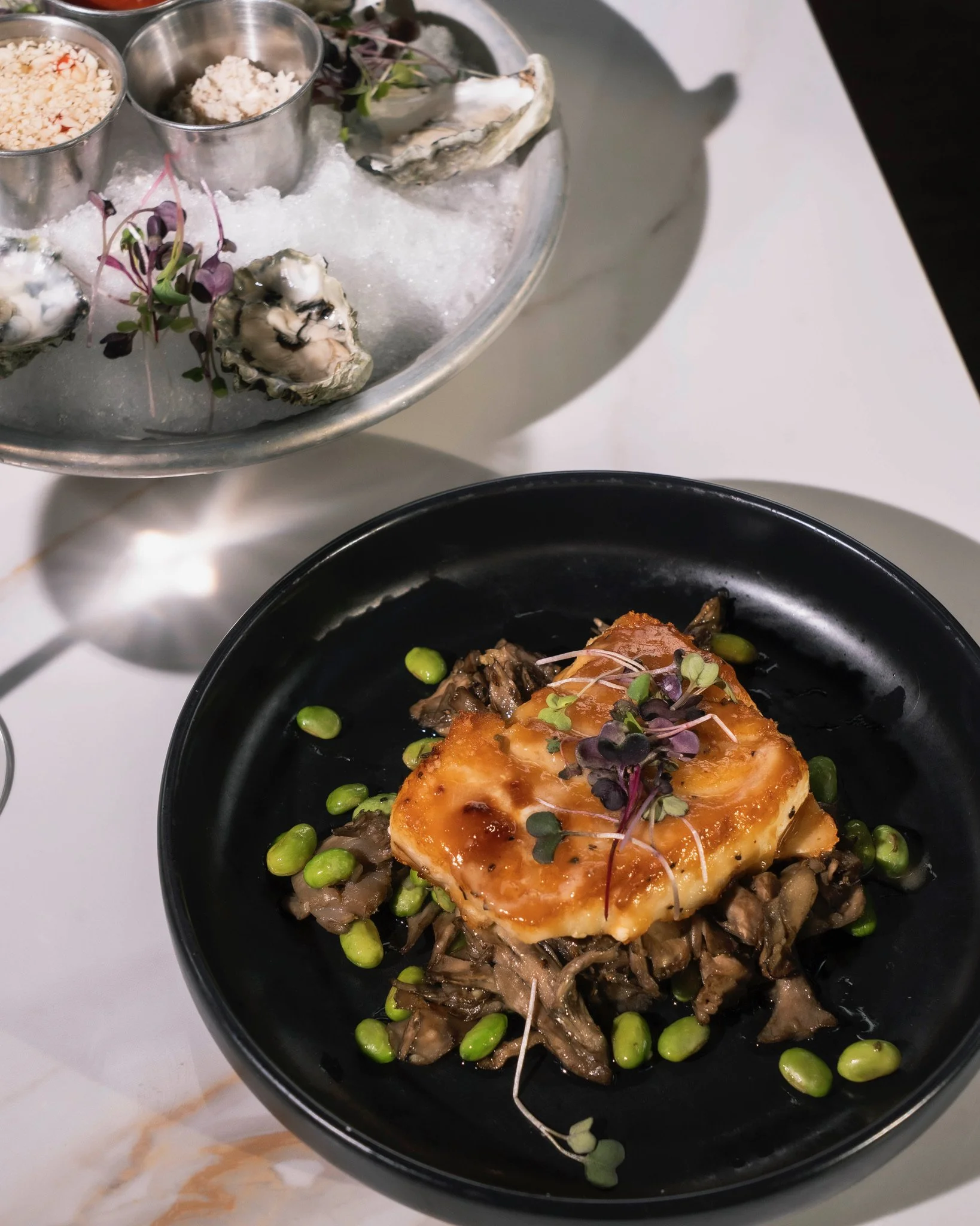 A dish with a piece of seared fish topped with a glaze, served over sautéed mushrooms and garnished with microgreens and edamame beans, with a seafood platter in the background.