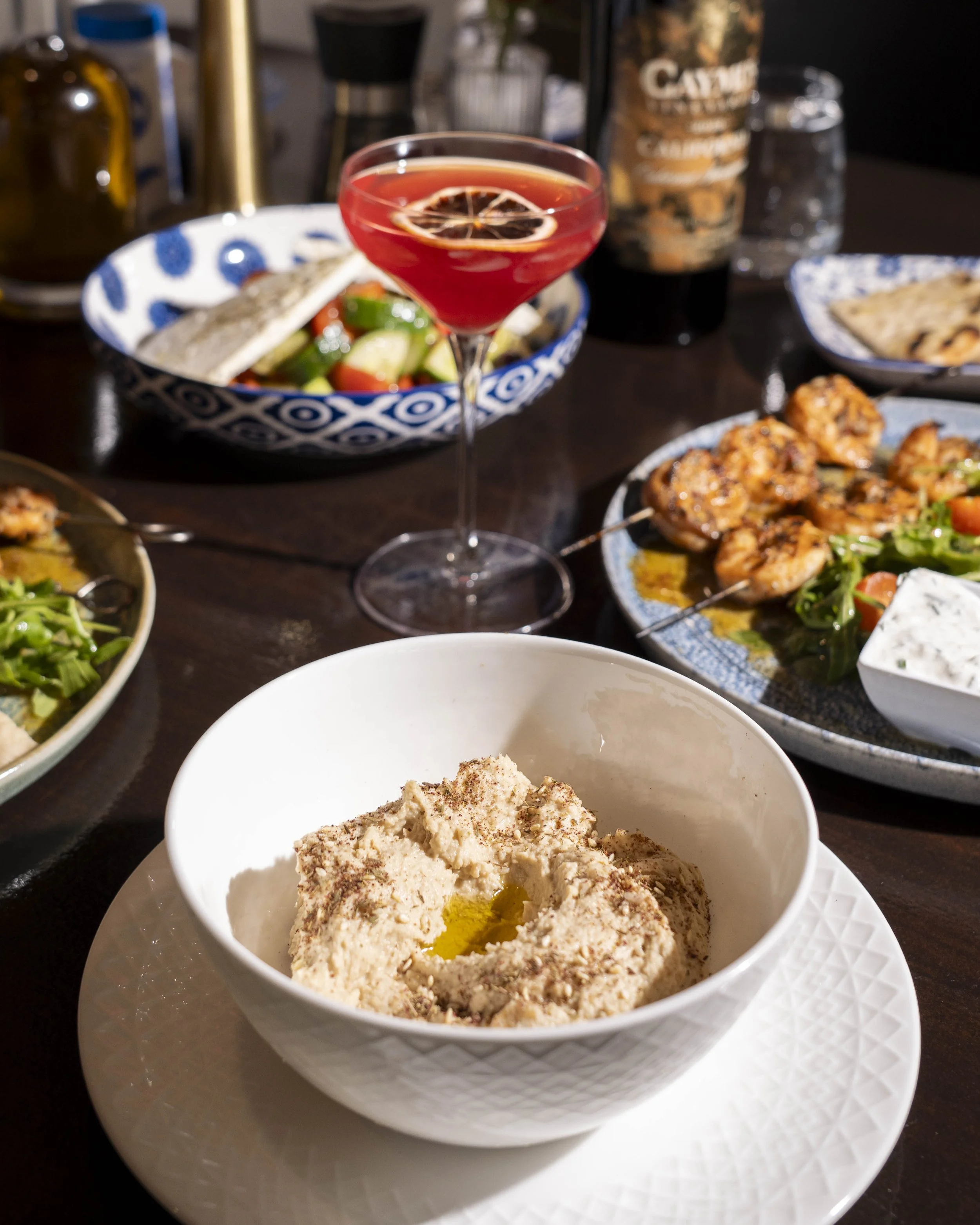 A dining table with various Middle Eastern dishes, including hummus with olive oil, grilled chicken skewers, a cucumber and tomato salad, and a cocktail with lemon slices.