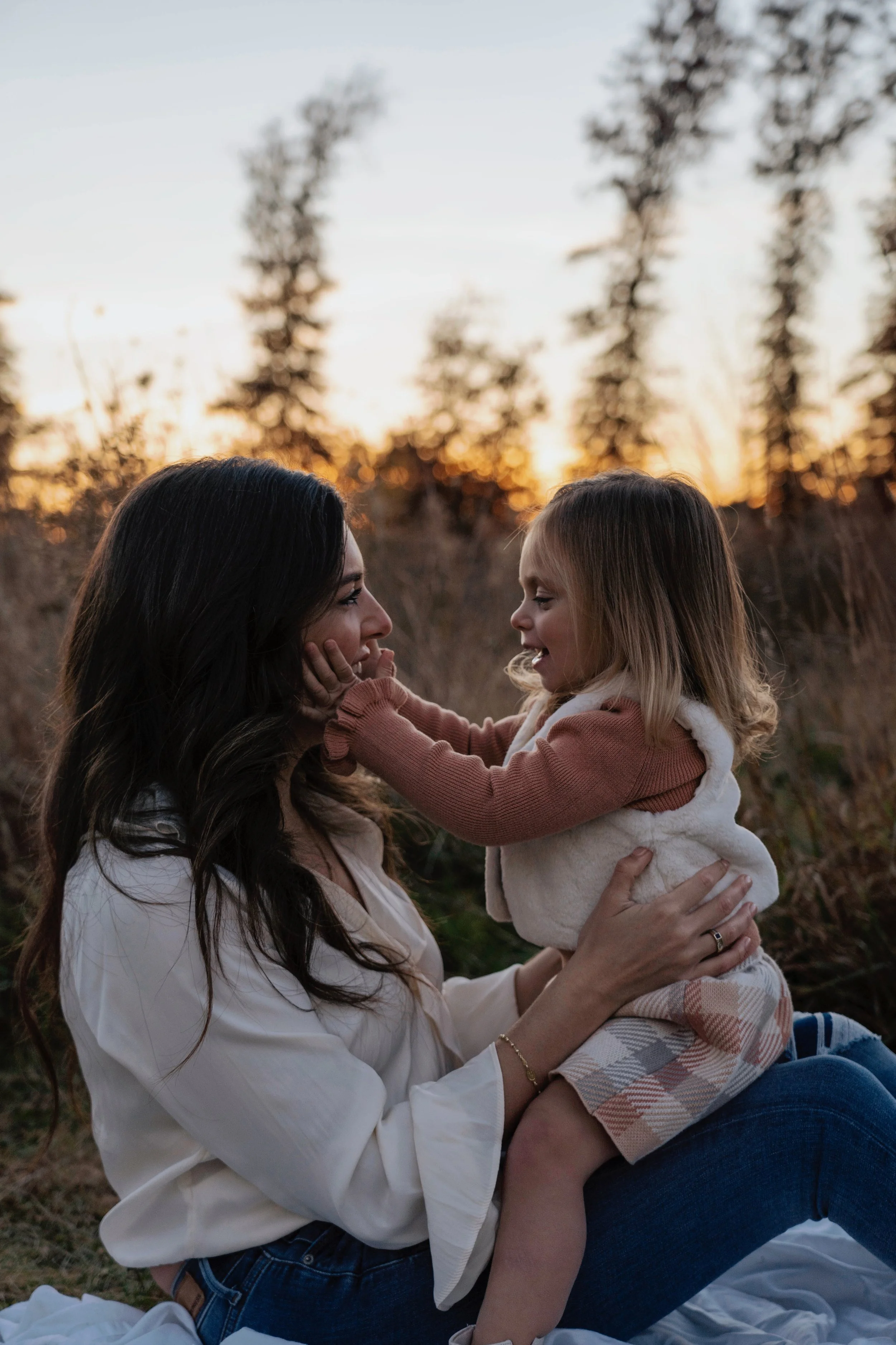 A woman and a young girl are sitting outdoors during sunset, smiling and playing near trees.