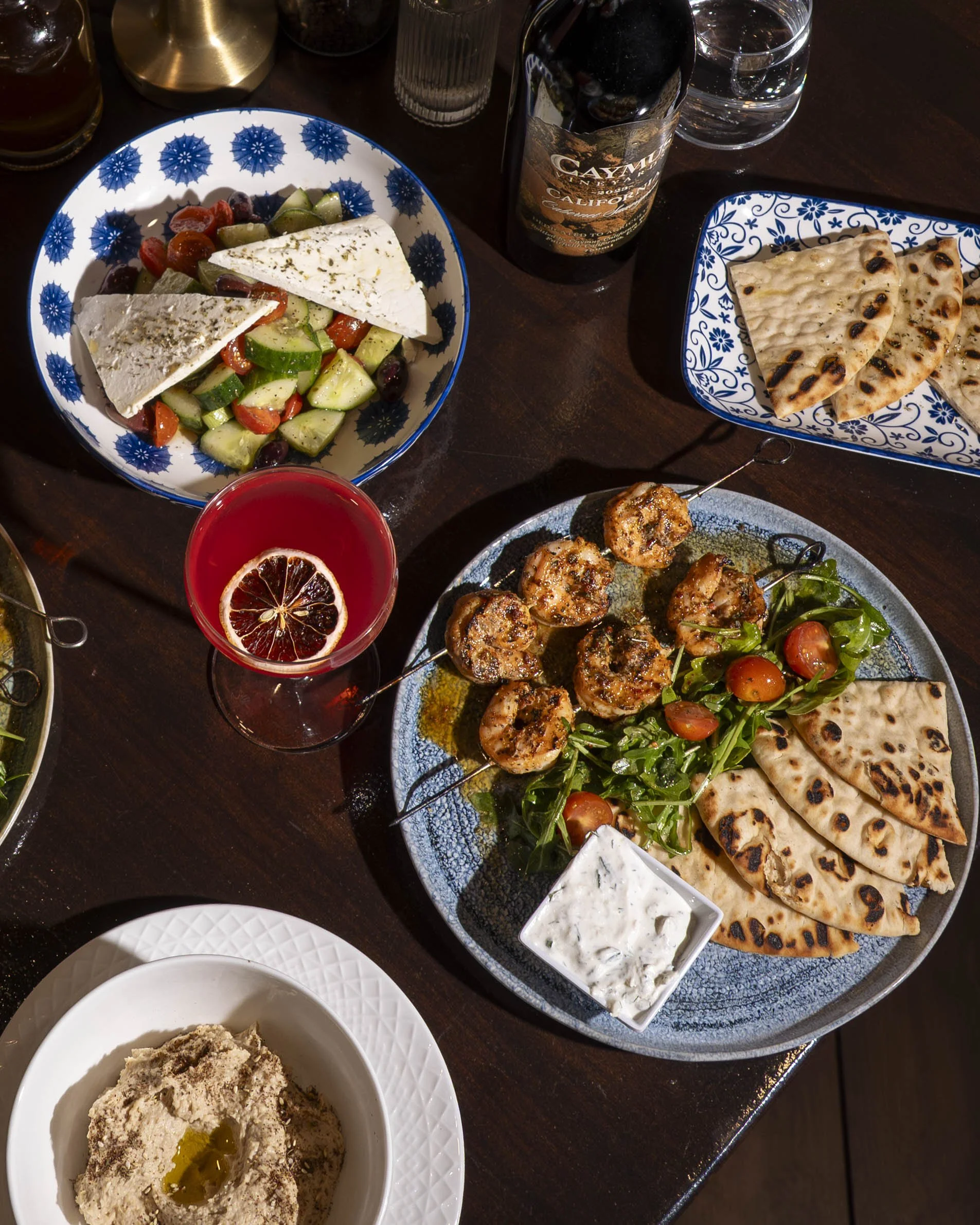 Table with Mediterranean food including grilled meat skewers, flatbread, salad with tomatoes and greens, feta cheese, a glass of red beverage with lemon slice, a bottle of Mexican beer, and a bowl of hummus.