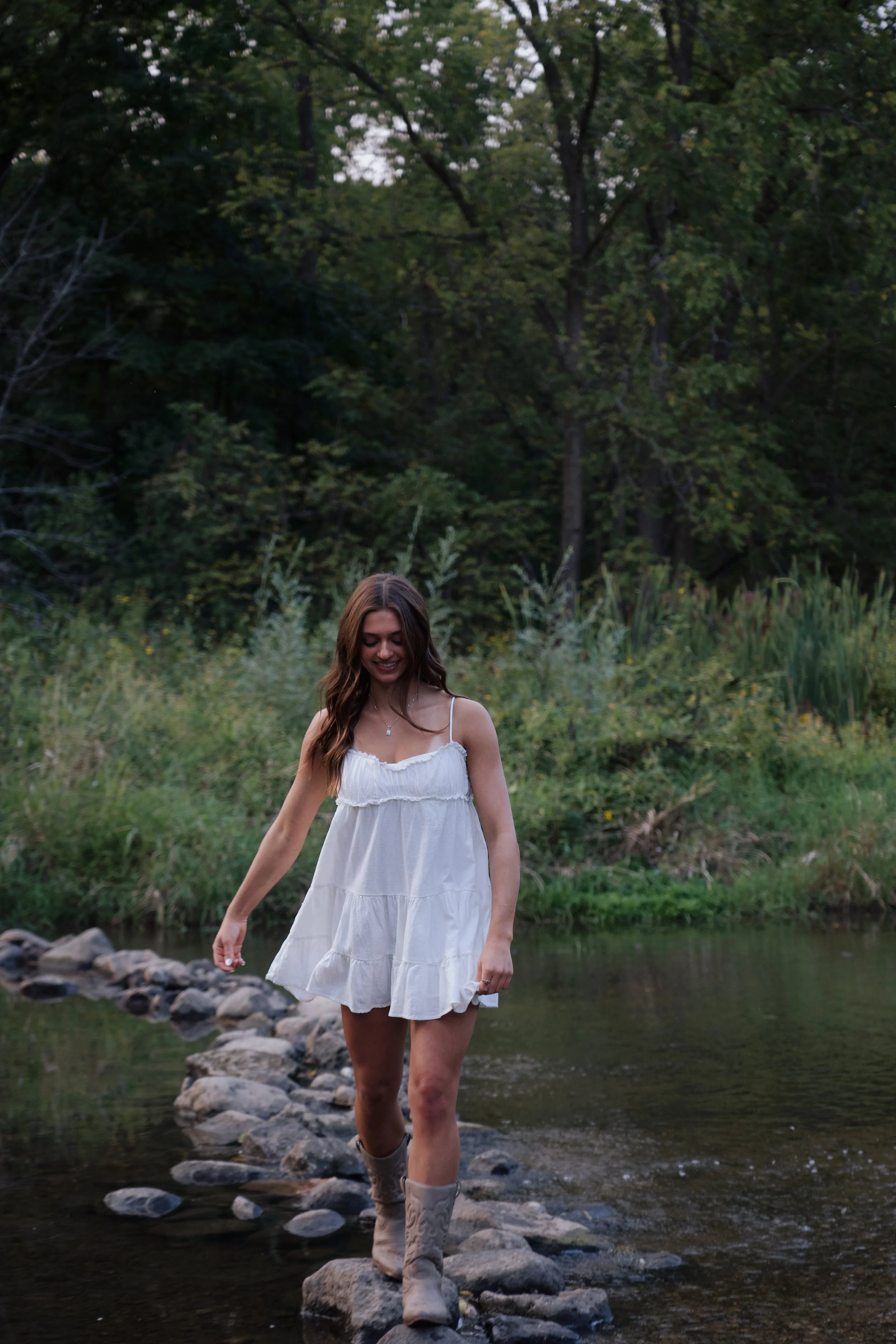 A young woman with long brown hair, wearing a white sundress and cowboy boots, walking across rocks in a shallow creek surrounded by green trees and bushes.