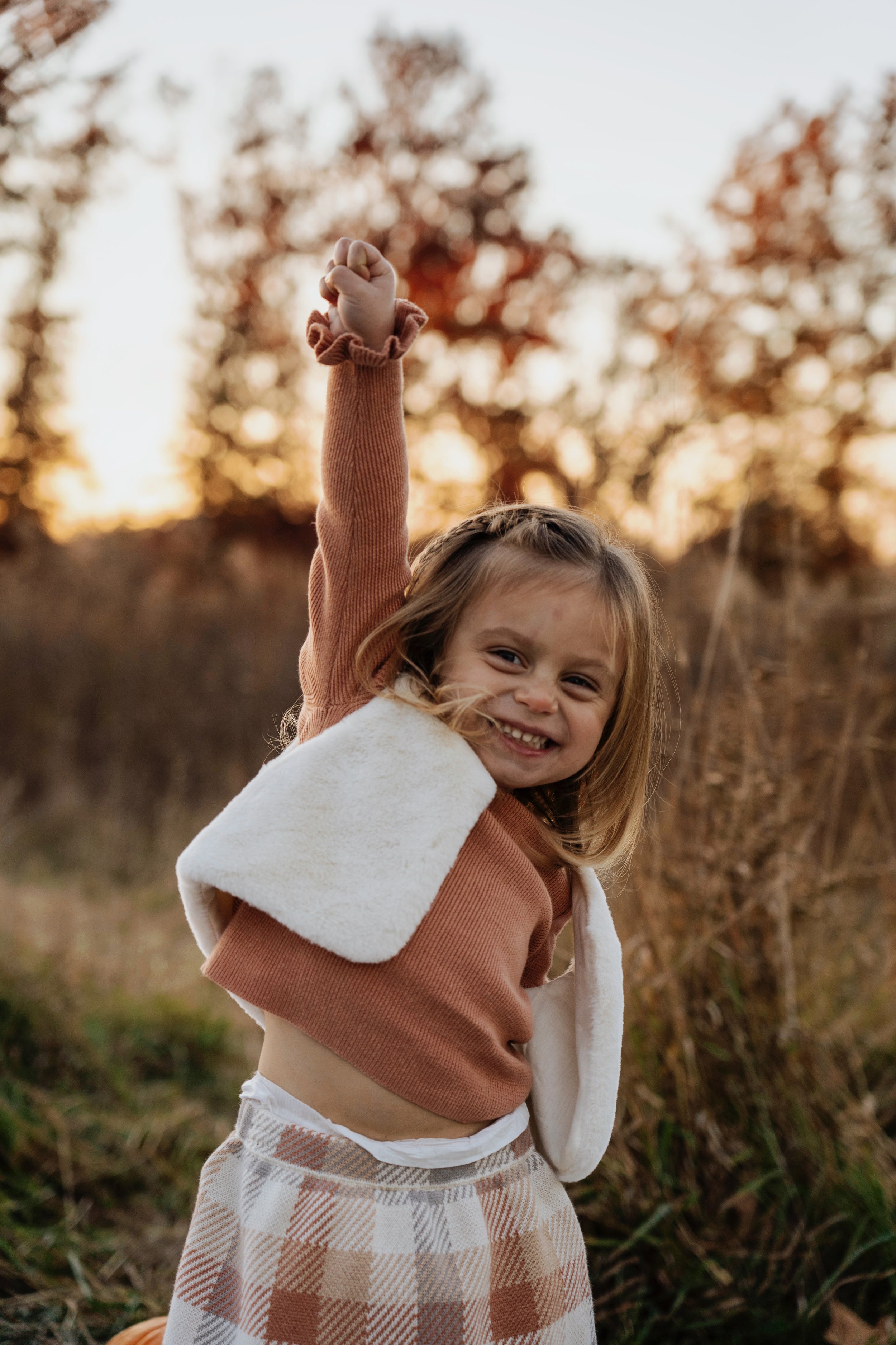 A young girl smiling and posing with one arm raised outdoors during sunset, wearing a brown sweater, a white vest, and beige plaid pants.