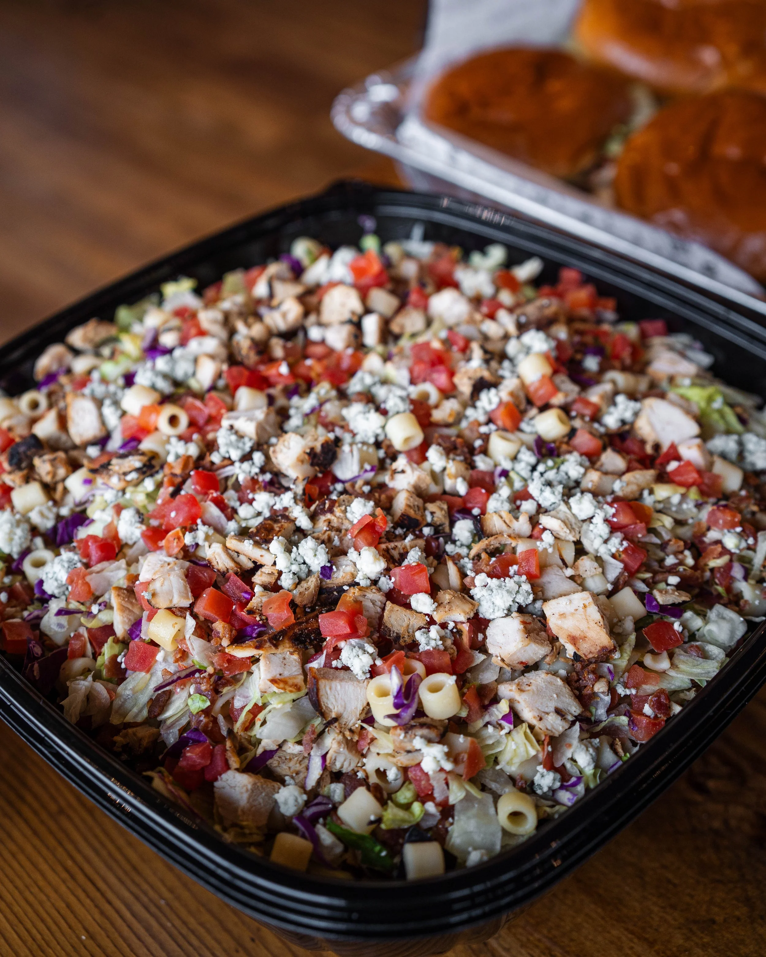 A large tray of chopped grilled chicken, pasta, tomatoes, and crumbled cheese on a wooden table.