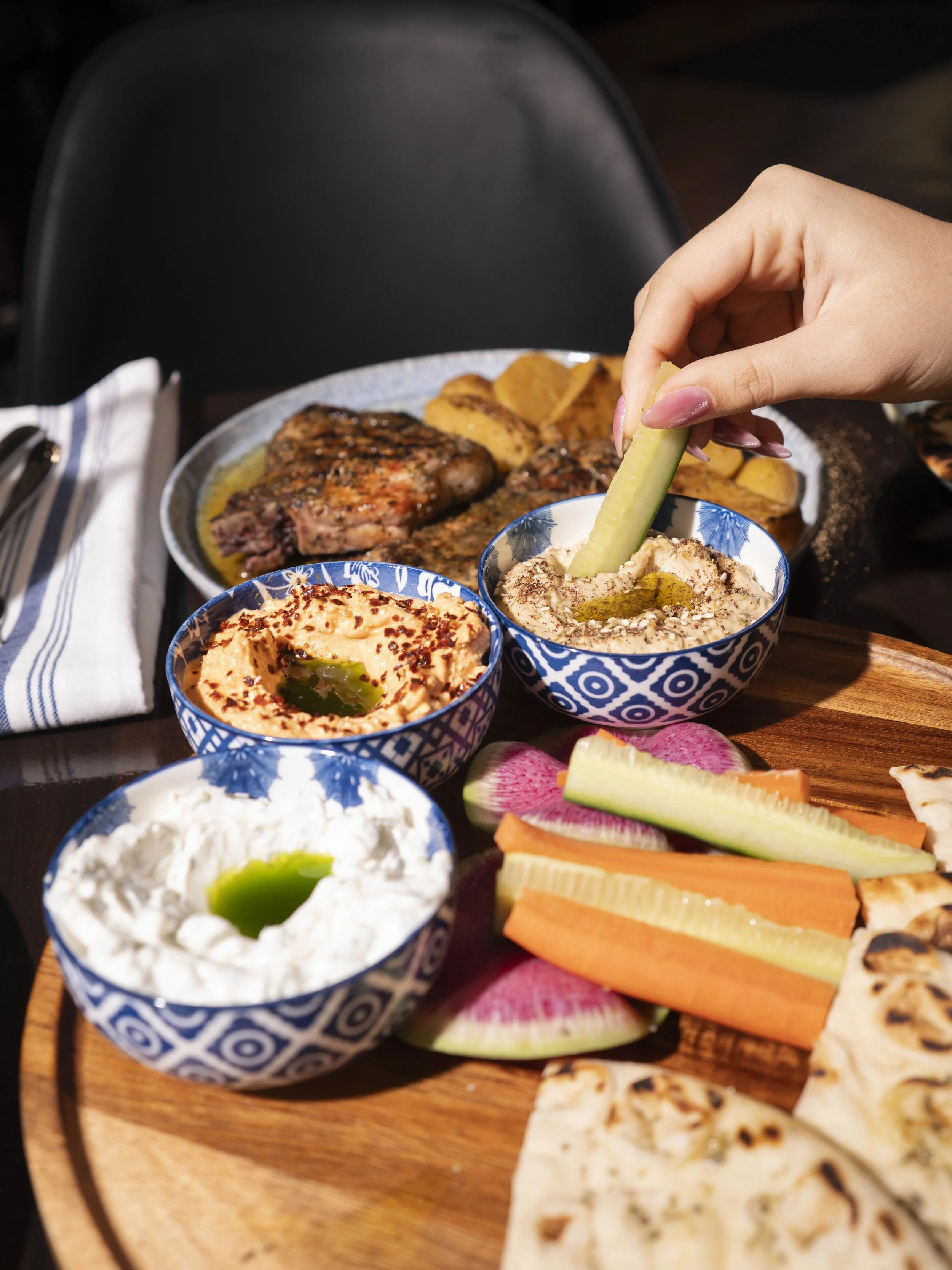 A hand dips a cucumber into three bowls of hummus. The bowls are on a wooden serving board with radishes, carrots, pita bread, roasted meat, and potato wedges.